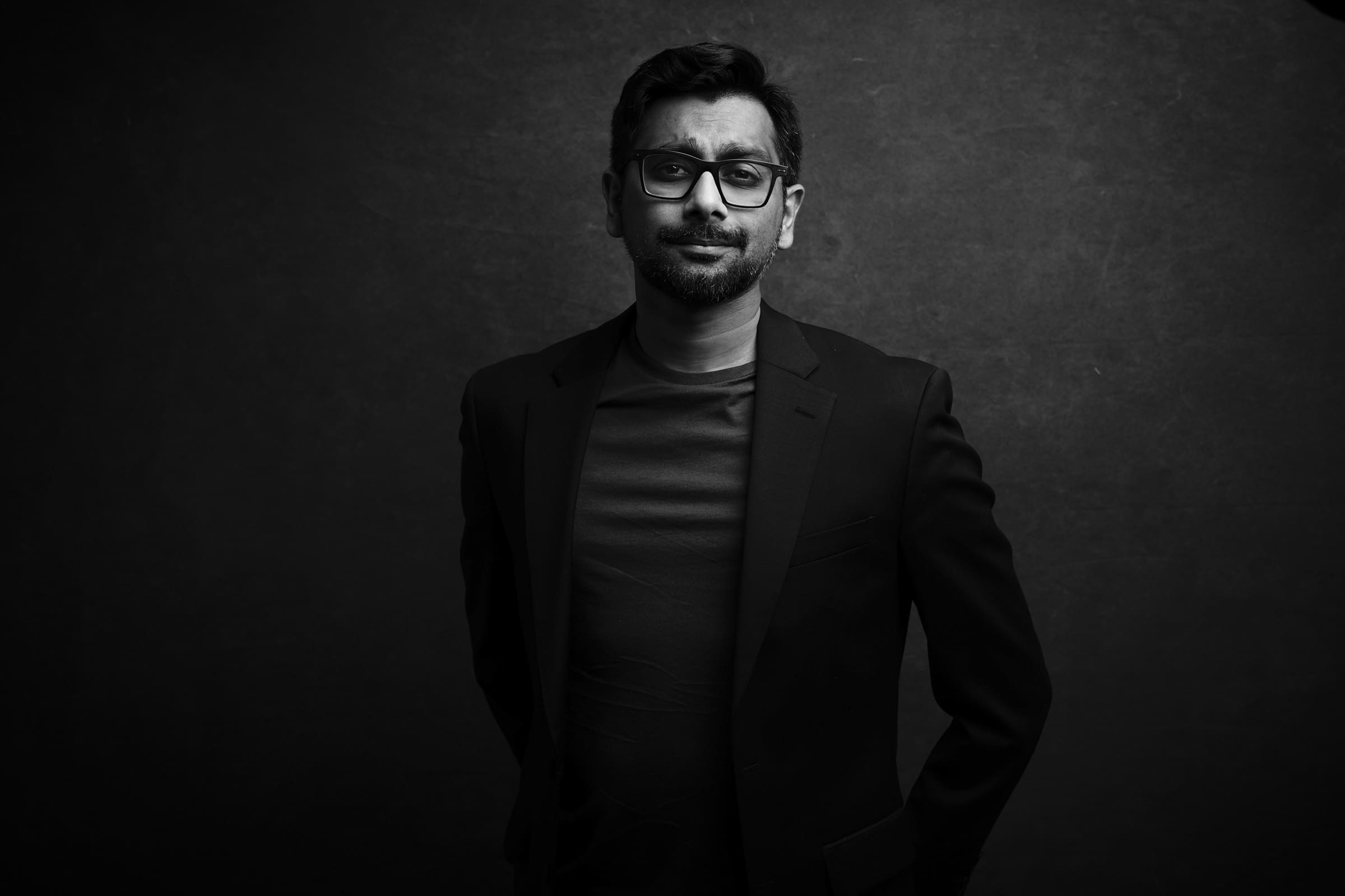 Headshot & branding session for dj rang A grayscale headshot captures a man with glasses and facial hair, wearing a dark blazer over a T-shirt. He stands against a textured gray background, exuding subtle confidence with his smile and hands behind his back—a perfect fit for any branding session.