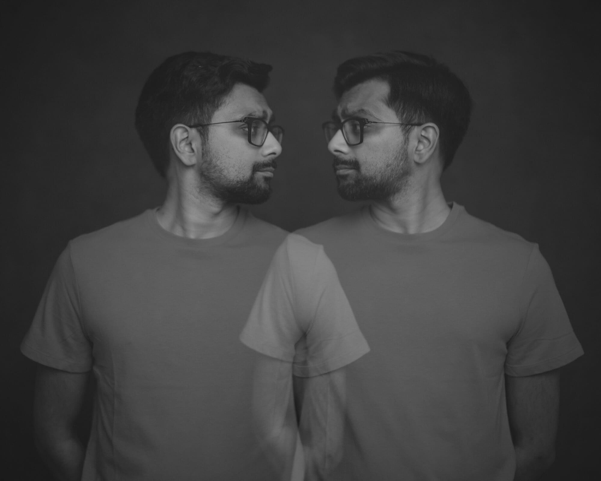 Headshot & branding session for dj rang A black and white headshot of a man in glasses and a plain t-shirt, with a double exposure effect revealing him contemplating his mirrored self. Perfect for a unique branding session, the neutral, blurred background enhances its introspective allure.