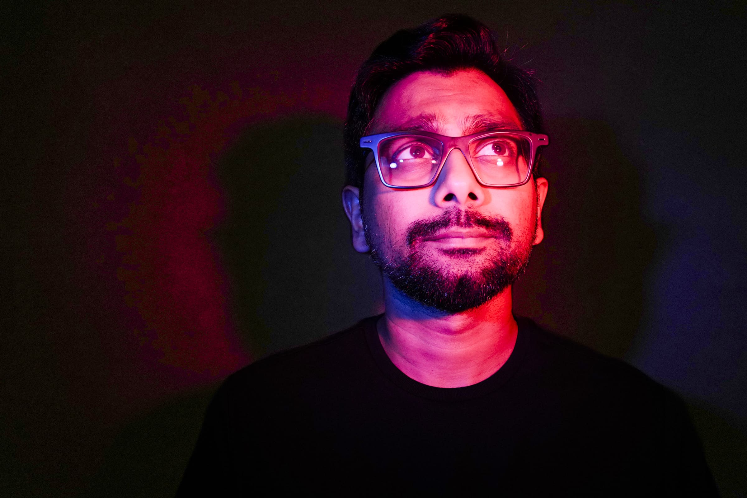 Headshot & branding session for dj rang A person with glasses looks up, captured in a striking headshot, illuminated by dramatic red and purple lighting against a dark background.