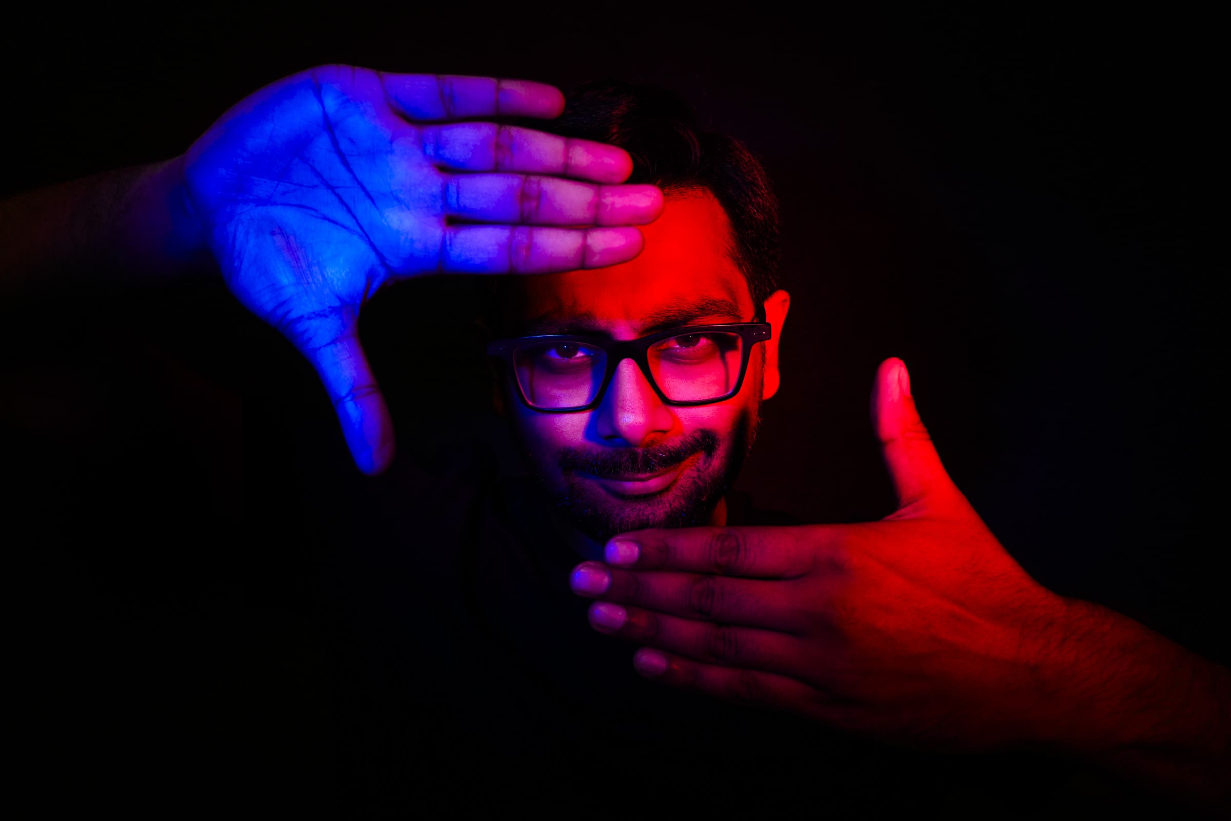 During a branding session, a person wearing glasses holds their hands in a rectangular shape in front of their face. One hand is bathed in blue light and the other in red, creating a striking contrast against the dark background, perfect for an impactful headshot as they look directly at the camera.