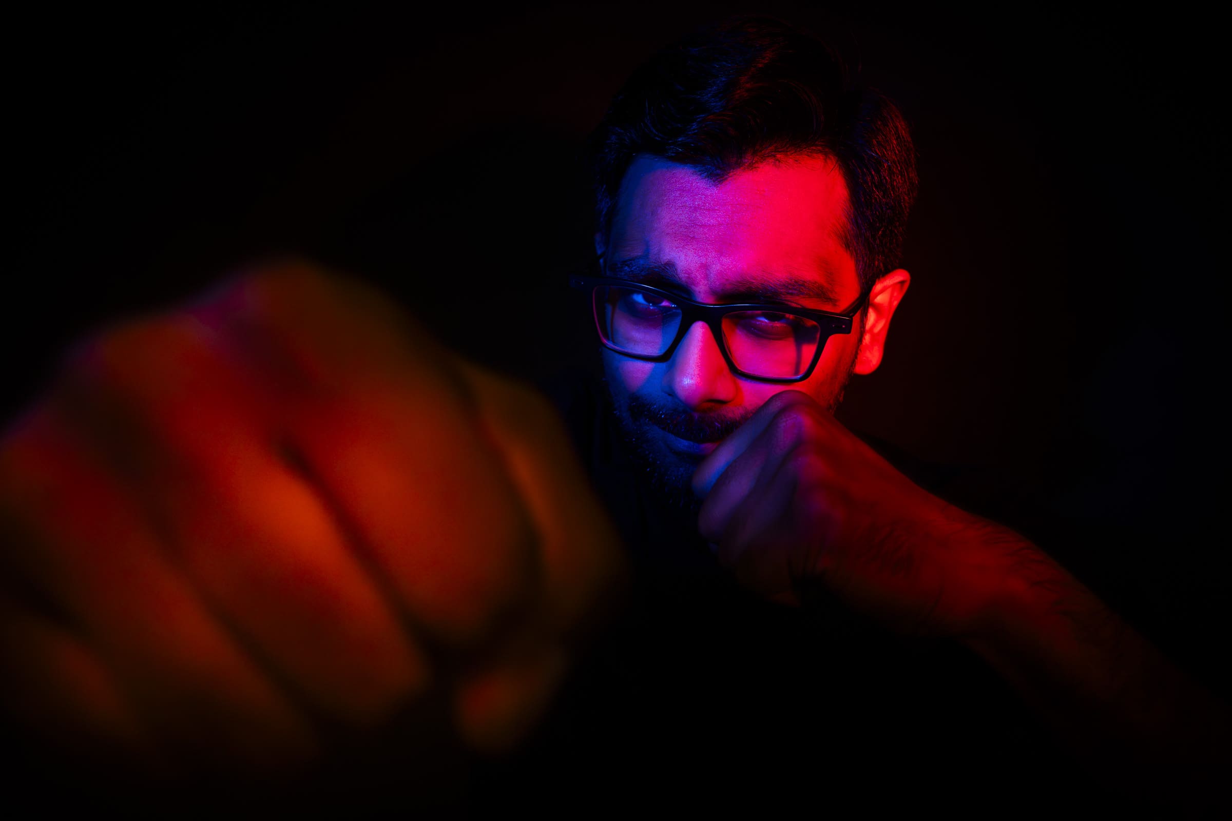 Headshot & branding session for dj rang In a striking headshot, a person wearing glasses assumes a boxing stance, one fist extended towards the camera. Dramatic red and blue lighting creates a high-contrast effect on their face and fists, perfect for an eye-catching branding session.