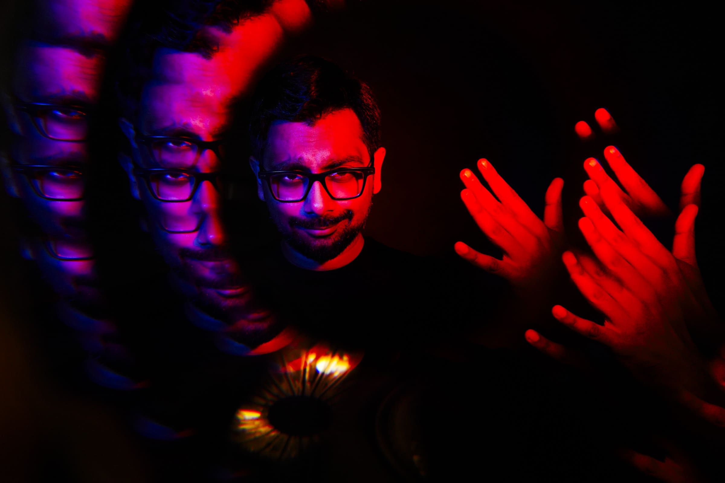 Headshot & branding session for dj rang A man with glasses is enveloped by mirrored reflections of his face and red-tinted hands against a dark backdrop. The image, reminiscent of a headshot, boasts a kaleidoscopic effect with vibrant colors, delivering an abstract composition ideal for a creative branding session.