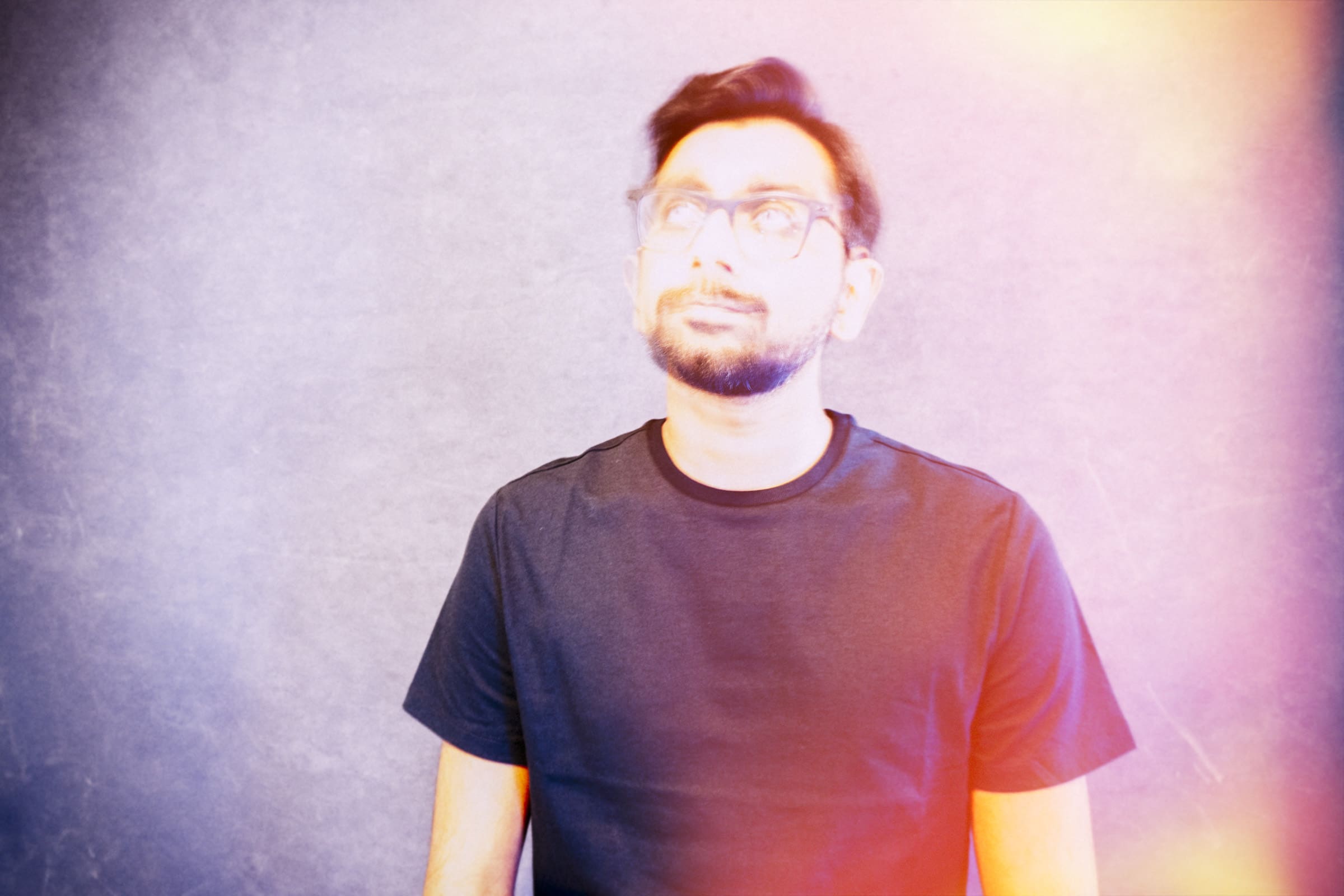 Headshot & branding session for dj rang A man with glasses and a beard wearing a black T-shirt stands against a gray background in this striking headshot. The image, perfect for a branding session, features colorful light leaks that add a vibrant effect.
