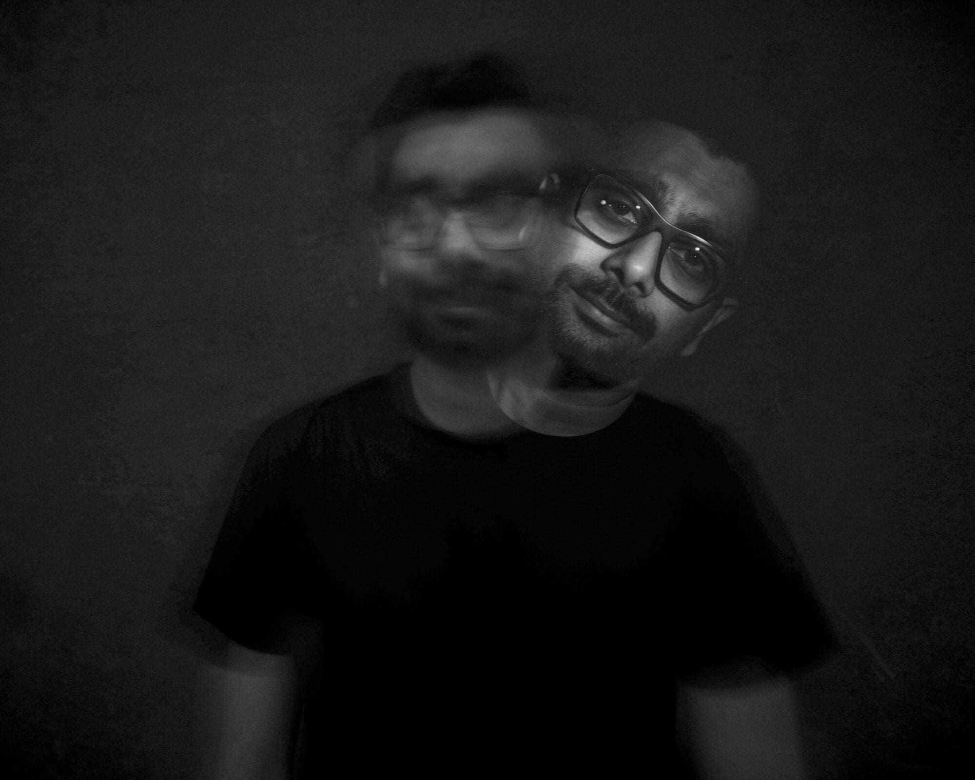 Headshot & branding session for dj rang A black and white headshot captures a man wearing glasses and a black shirt. The image features a motion blur effect, creating two overlapping impressions of his face, perfect for a dynamic branding session. The background is dark and plain, enhancing the striking visual impact.