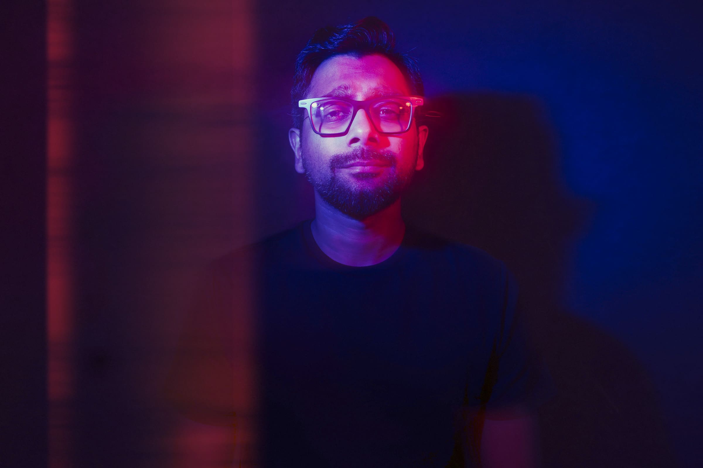 Headshot & branding session for dj rang A man with glasses stands against a dark background, illuminated by dramatic red and blue lighting. In this headshot, shadows and light create a striking contrast, highlighting the contemplative expression on his face—ideal for a powerful branding session.