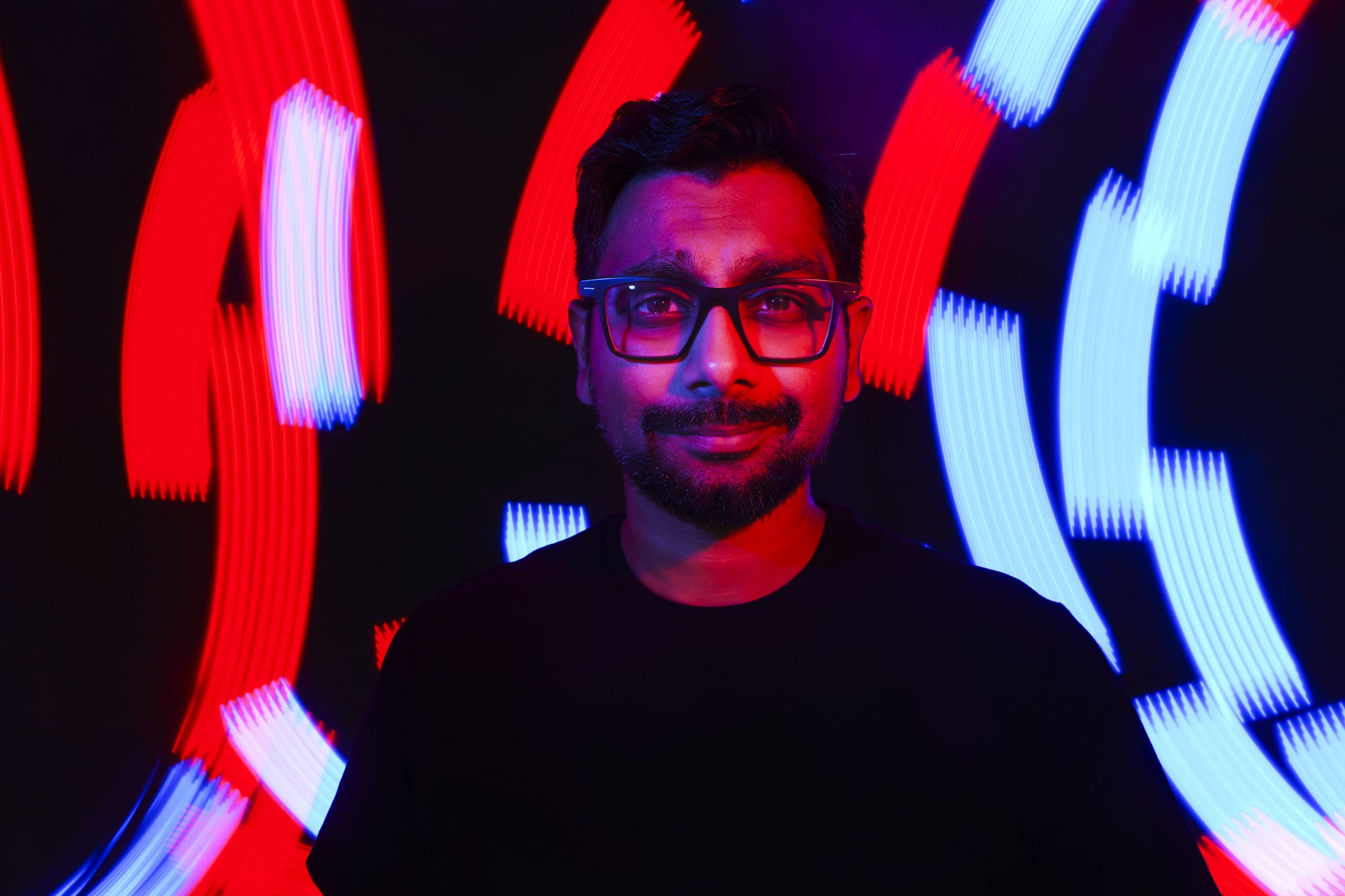 Headshot & branding session for dj rang A man with glasses poses for a headshot against a background of red and blue light streaks. Wearing a dark shirt, he brings an intriguing dynamic to this branding session as the vibrant lights create a colorful pattern around him.