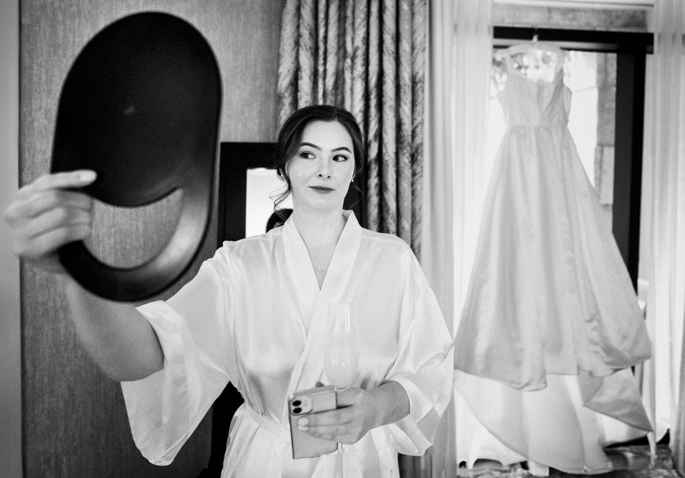 A woman in a white robe holds a handheld mirror and a drink, standing near large windows with curtains in a room at the Umstead Hotel. A hanging wedding dress graces the background, adding elegance to this black and white scene.
