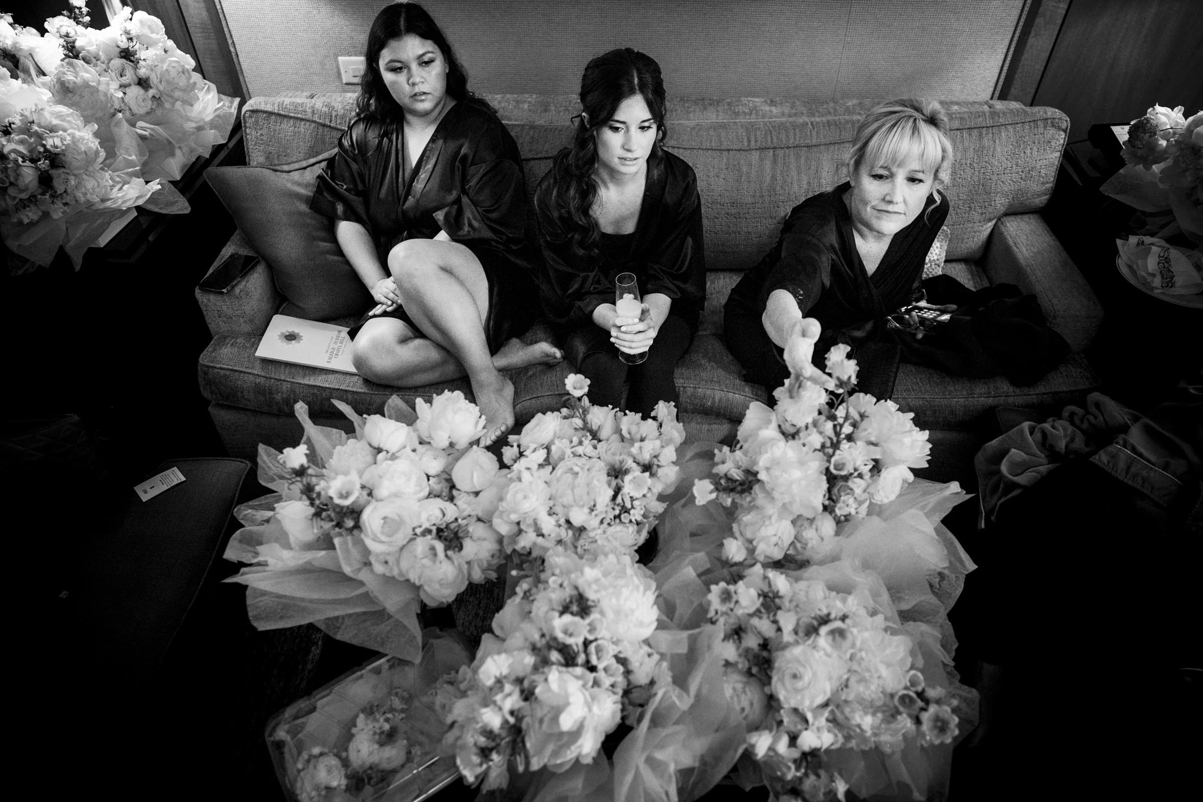 Three women in robes sit on a sofa surrounded by bouquets of flowers at an Umstead Hotel wedding. One holds a glass, while another reaches towards a bouquet. The setting is cozy and intimate, with a focus on floral arrangements. The image is in black and white.