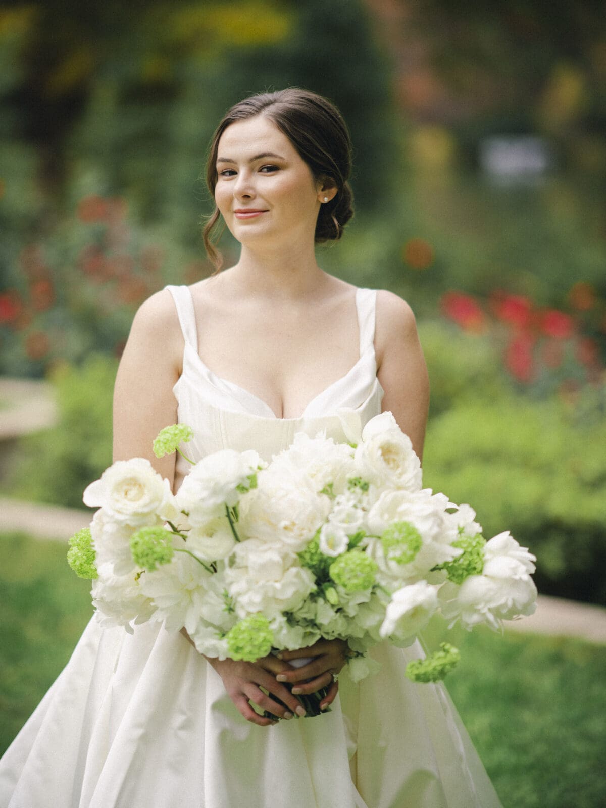 A bride in a white dress holds a large bouquet of white and green flowers, embodying the elegance of an Umstead Hotel Wedding. She stands outdoors with a blurred, colorful garden background, the greenery and flowers around her adding to the serene atmosphere.