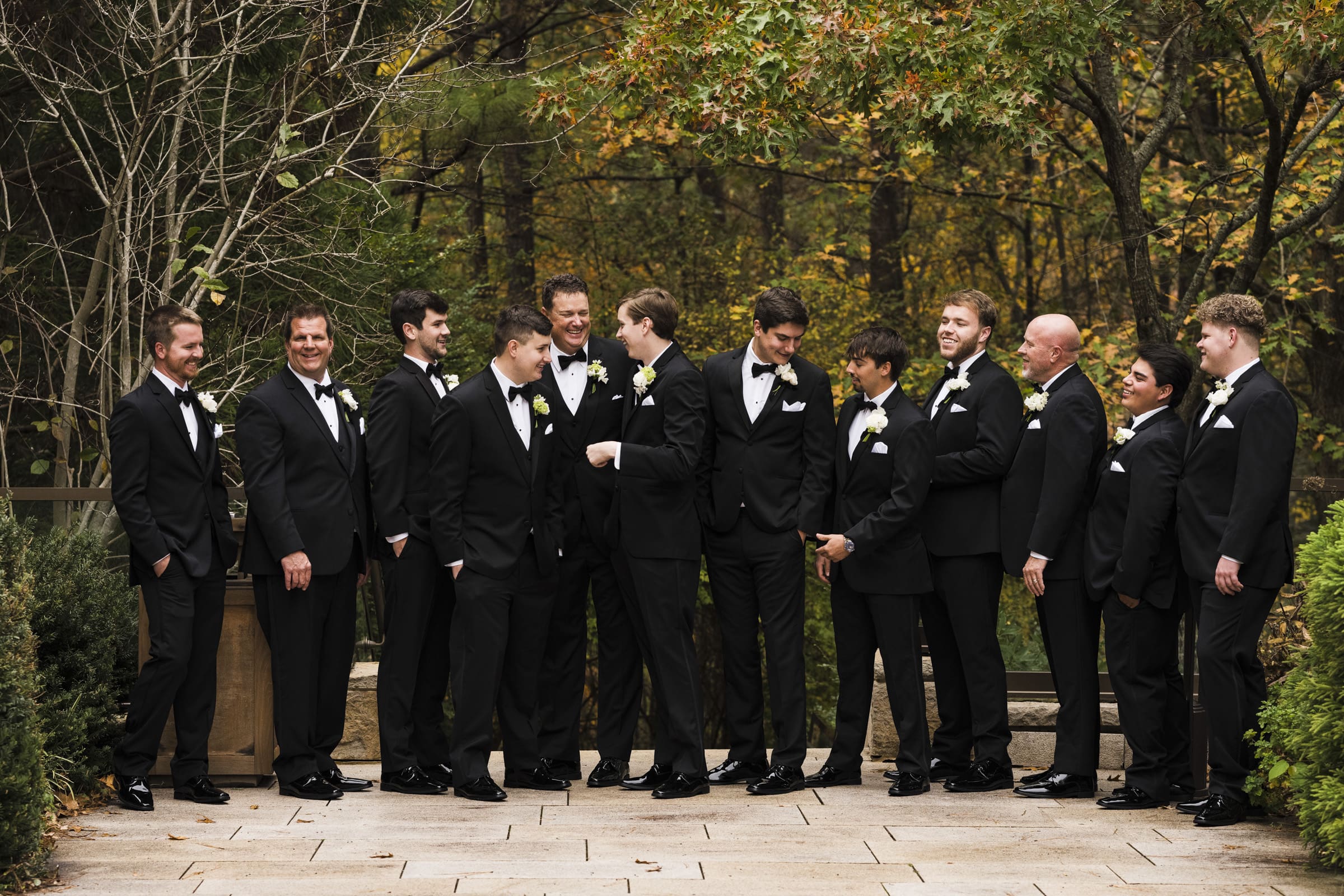 A group of men dressed in black tuxedos with bow ties stand outdoors on a stone path surrounded by greenery, embodying the charm of an Umstead Hotel wedding. They are all smiling and appear to be in a celebratory mood, possibly for a wedding or formal event.