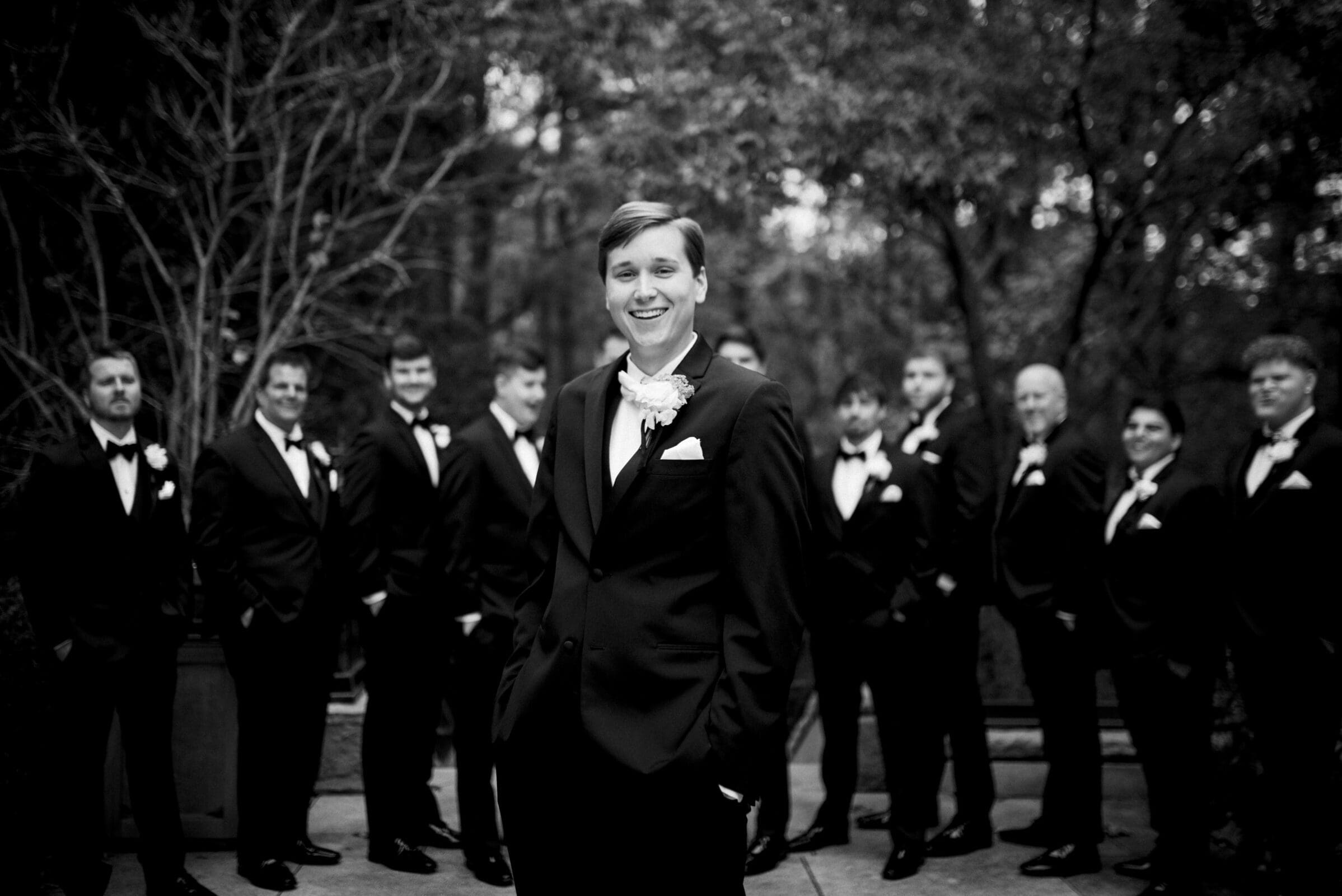 A black and white photo captures the groom at an Umstead Hotel Wedding, smiling with hands in pockets. He stands confidently in front of his groomsmen, who are also dressed in suits, arranged casually against a backdrop of trees.