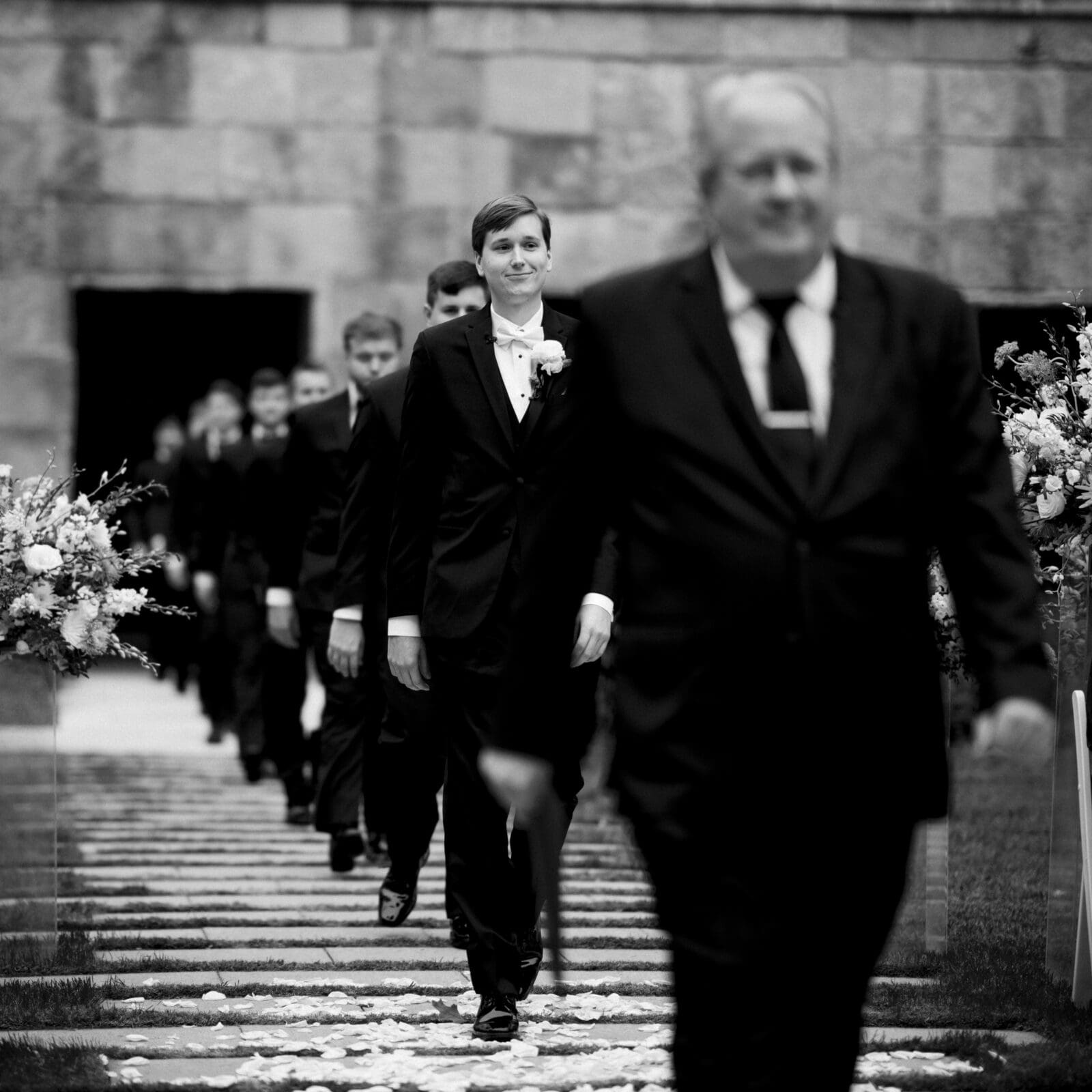 A group of people in formal attire walks in a line on the stone pathway at an elegant Umstead Hotel wedding. The focus is on a person smiling, wearing a suit and boutonniere. Flower arrangements line the path, leading to a grand stone building. The image is captured in black and white.