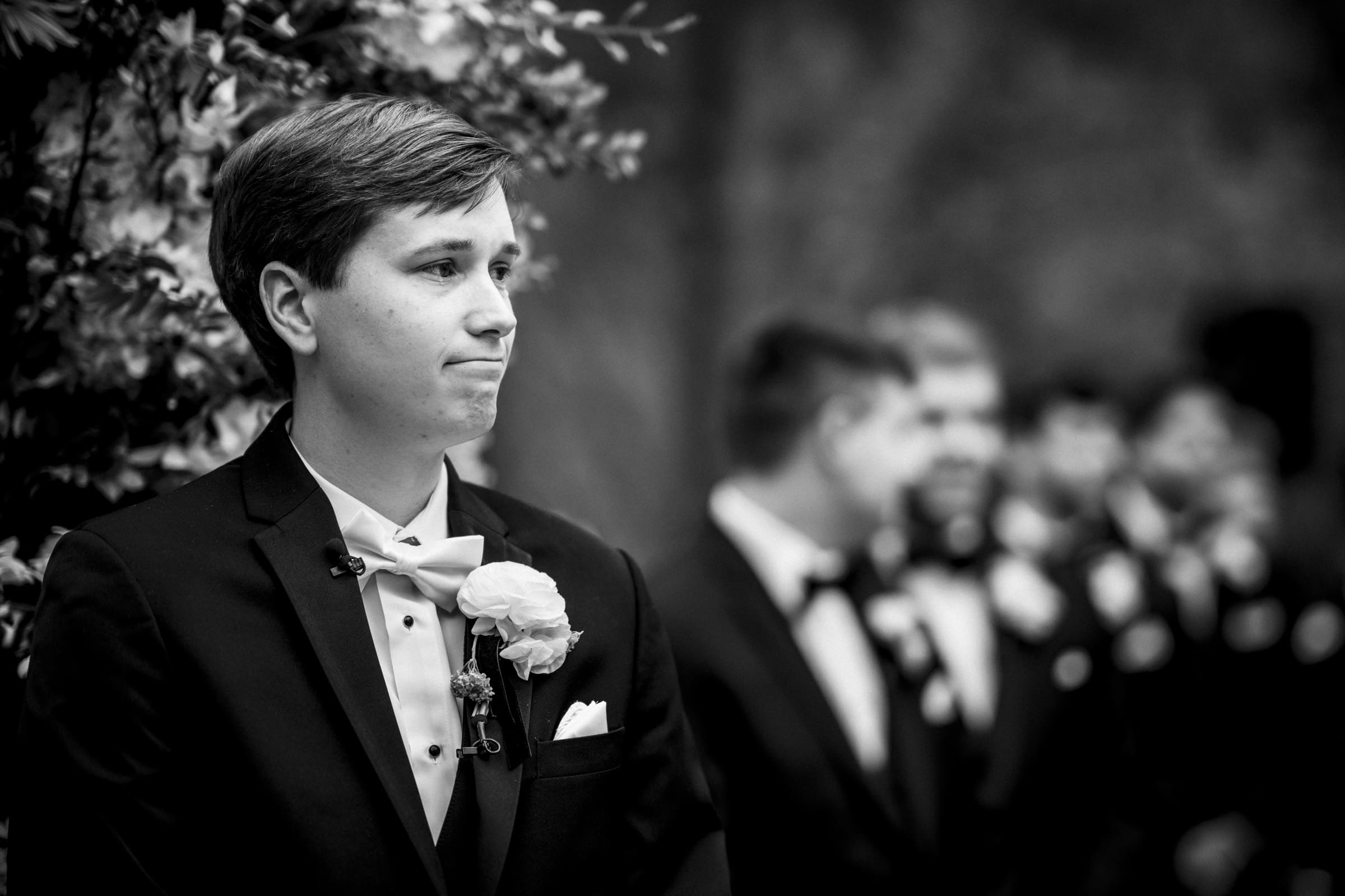 In a black and white image from an Umstead Hotel Wedding, the groom, adorned with a boutonniere on his tuxedo, stands at the altar looking slightly emotional. In the blurred background, groomsmen in formal attire stand in a line.