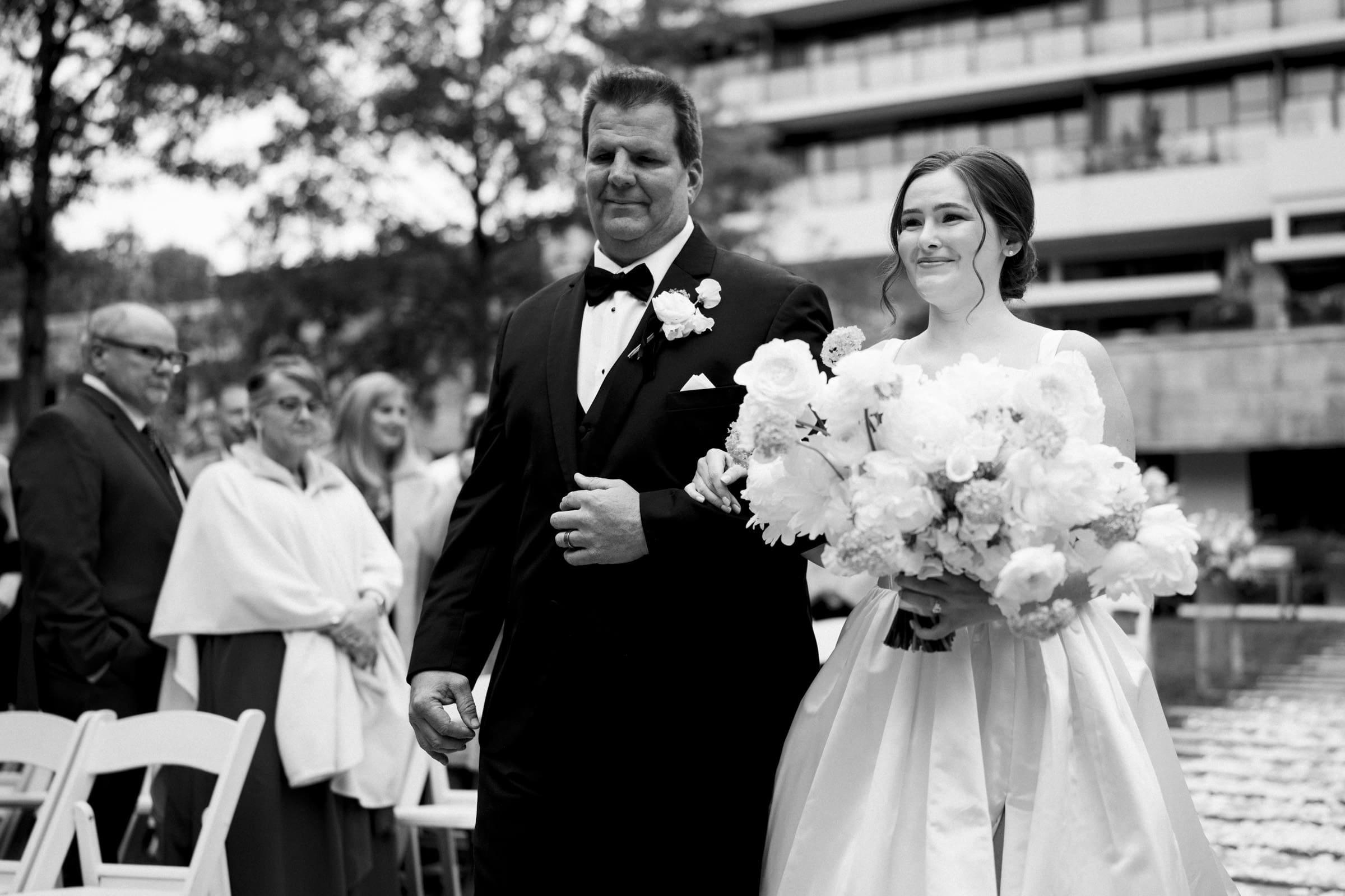 A bride in a white dress holds a bouquet of flowers as she walks down the aisle with an older man in a suit at an Umstead Hotel wedding. They are outdoors, with guests seated in the background. The image is captured beautifully in black and white.