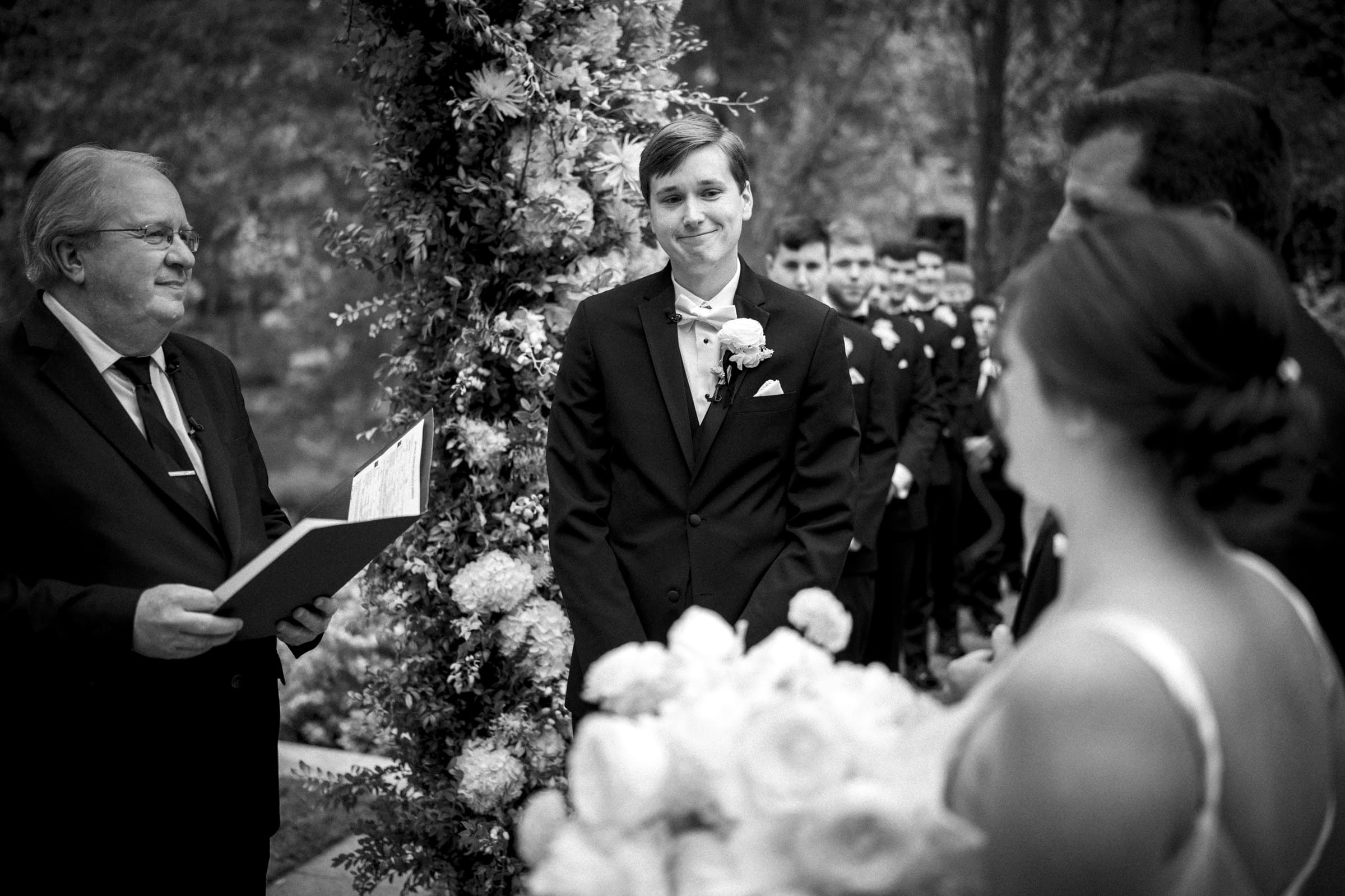A groom in a suit stands at an outdoor wedding ceremony, smiling at the bride. They are surrounded by flowers, with guests visible in the background. The officiant holds a book while conducting the ceremony. Captured in black and white, this Umstead Hotel Wedding exudes timeless elegance.