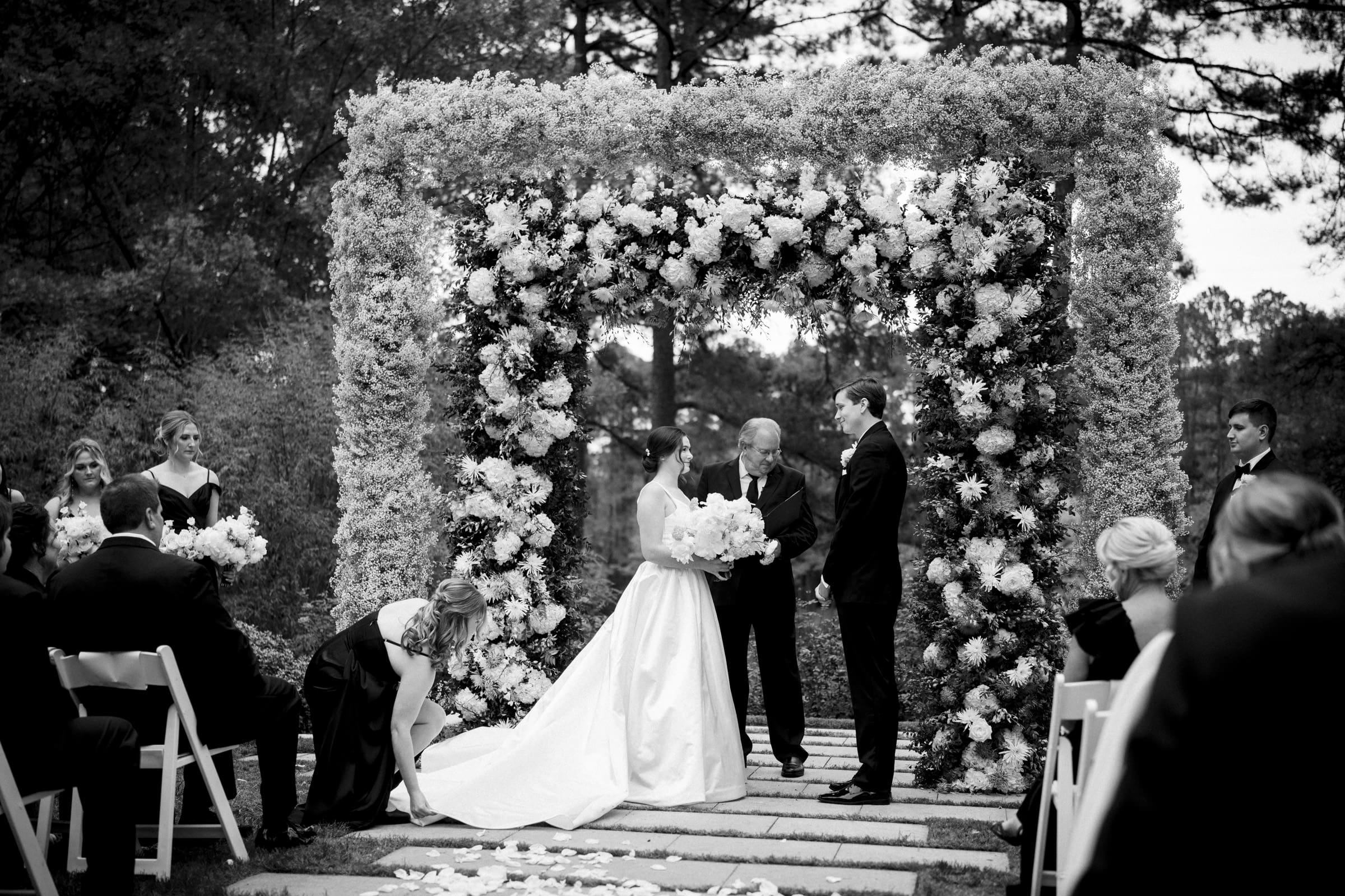 A black and white photo of an Umstead Hotel wedding captures the bride and groom under a floral arch, exchanging vows. Bridesmaids and groomsmen stand nearby, while a guest is seated in the foreground. One bridesmaid gently adjusts the bride's dress.