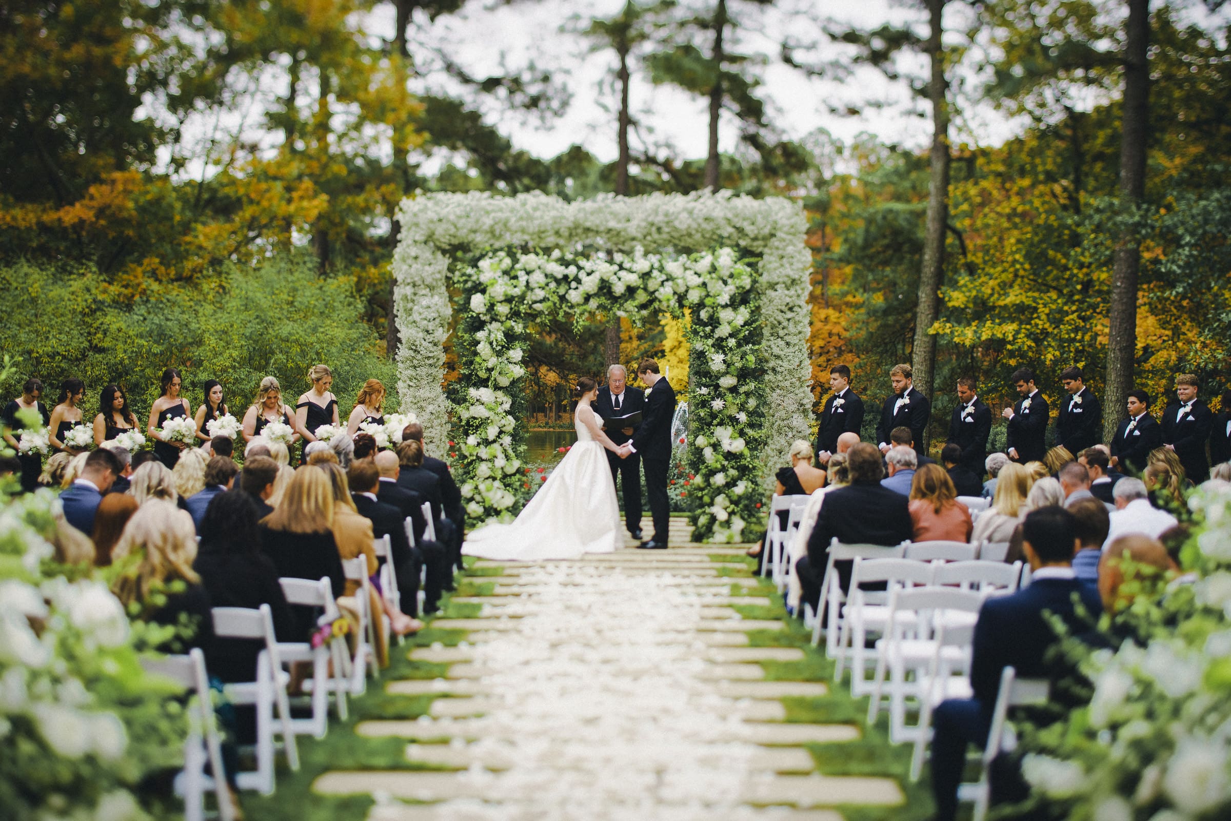 A bride and groom stand under a floral arch during a picturesque Umstead Hotel wedding. Guests are seated on either side of an aisle adorned with white petals, all set amidst trees showcasing vibrant green and autumn-colored leaves.