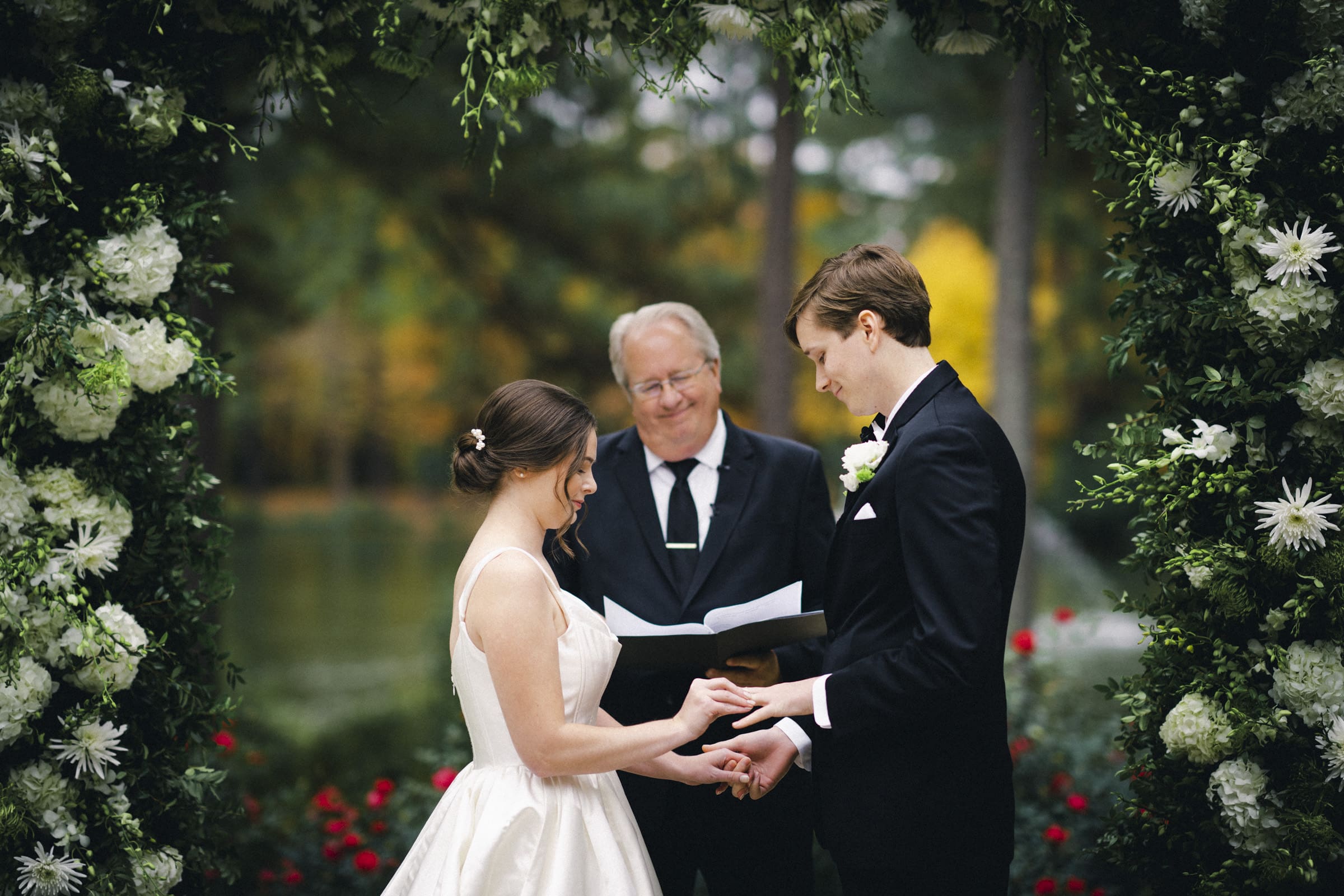 During their Umstead Hotel wedding, the couple exchanges rings beneath a floral arch. The bride, radiant in her white dress, and the groom, dashing in his black suit, are accompanied by a smiling officiant holding a booklet. Lush greenery and flowers beautifully frame the enchanting scene.