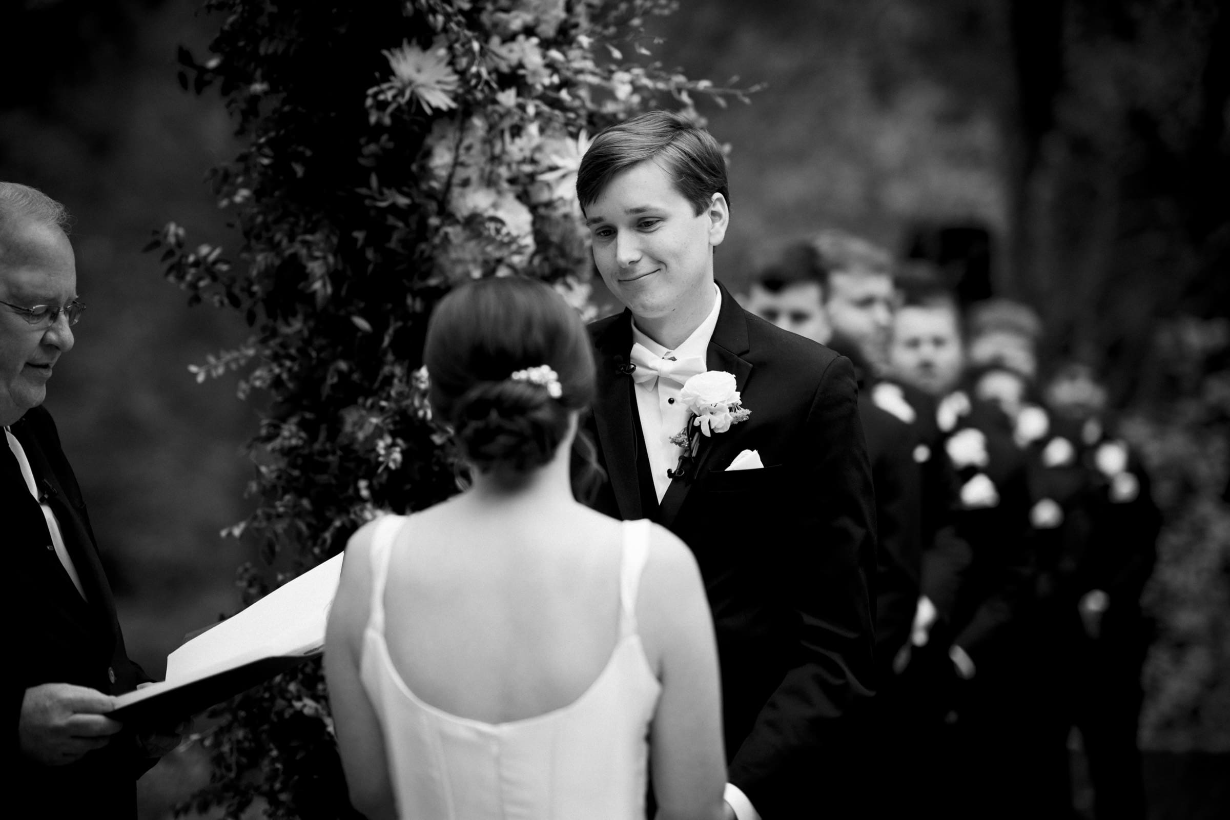 A black and white image captures an elegant Umstead Hotel wedding ceremony. A bride and groom stand facing each other under a floral arch, with the groom smiling. A group of men in matching suits line up in the background, while an officiant stands to the side with a book.