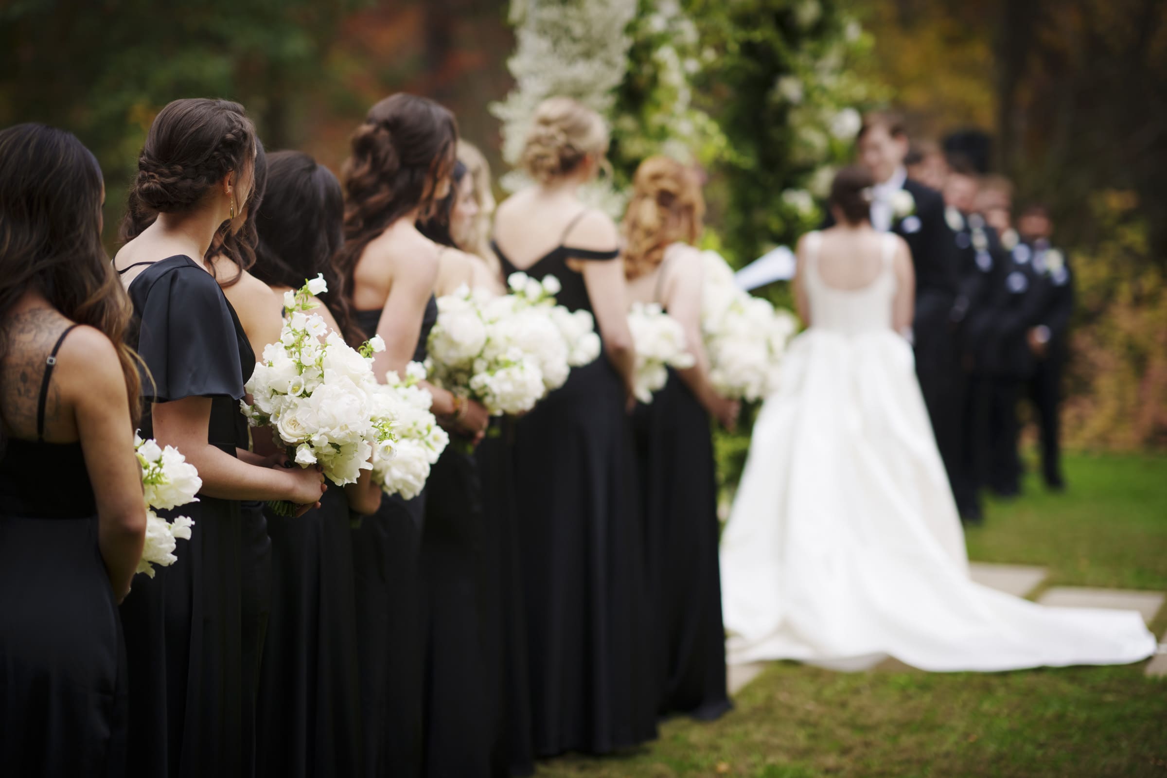 A bride in a white gown stands at an outdoor altar with a groom and officiant, capturing the elegance of an Umstead Hotel Wedding. Bridesmaids in black dresses, holding white bouquets, line up on the grass. Greenery and flowers adorn the backdrop, suggesting a wedding ceremony in progress.