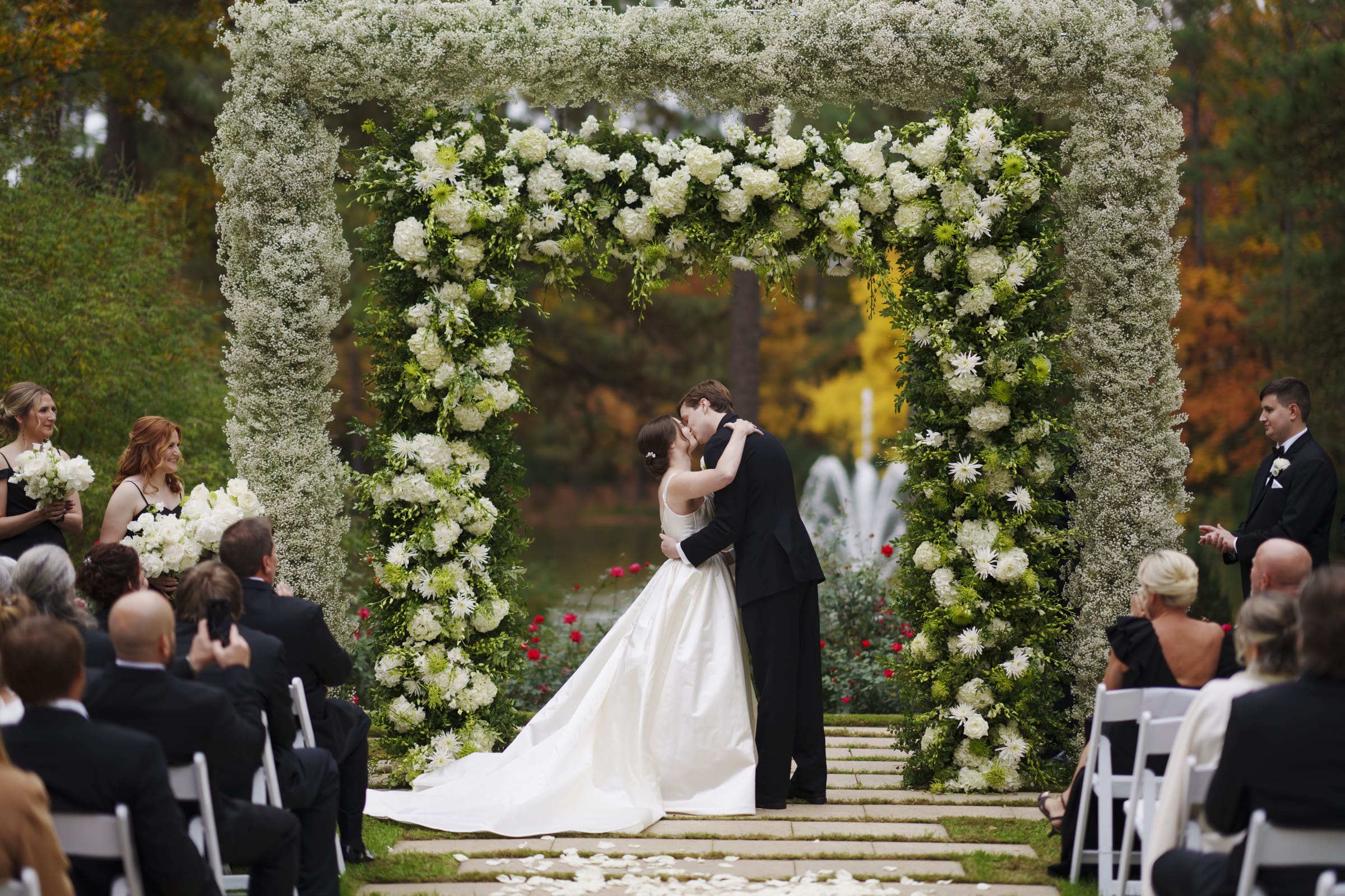 At their elegant Umstead Hotel wedding, a newlywed couple shares a kiss under a large, square-shaped floral arch adorned with white and green blooms. Surrounded by seated guests in formal attire, the outdoor setting features lush trees and the gentle sound of a nearby fountain.