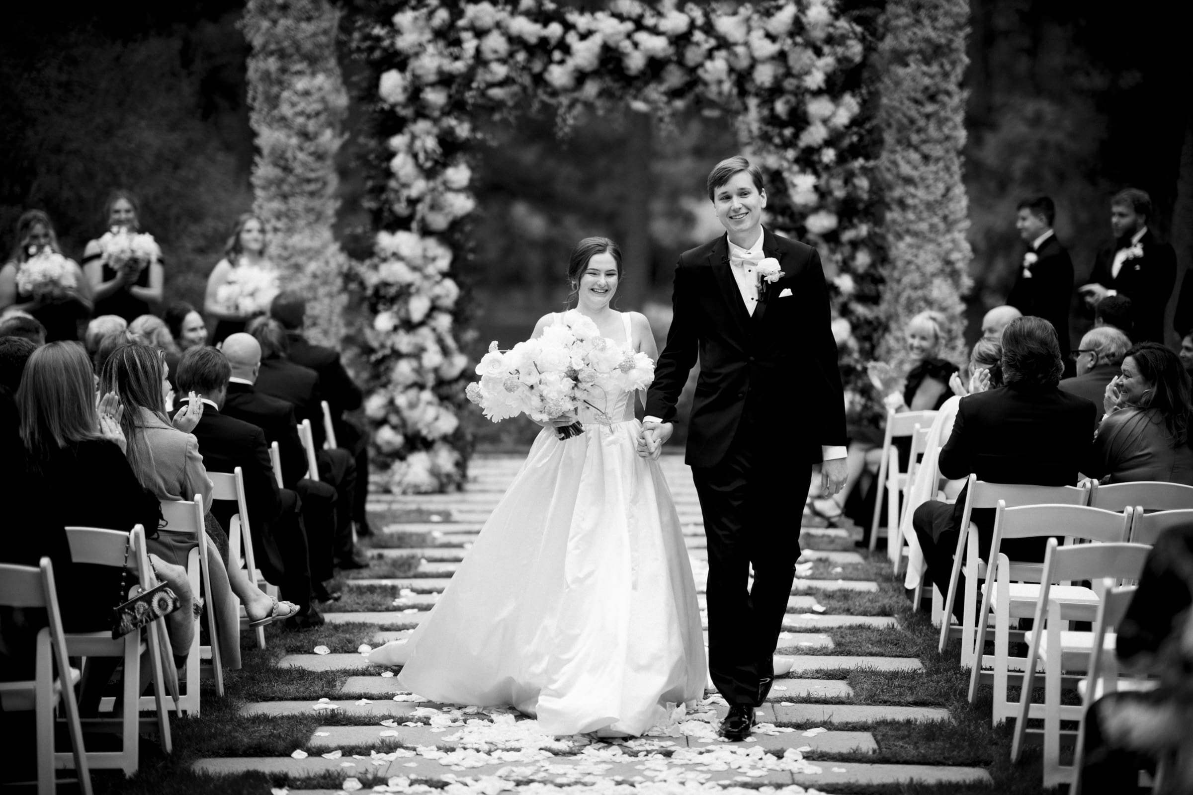 A bride and groom walk down an aisle lined with seated guests at their Umstead Hotel wedding. The groom wears a dark suit, while the bride stuns in a flowing white gown and holds a bouquet. Behind them, a floral arch adds to the joyful and celebratory atmosphere.