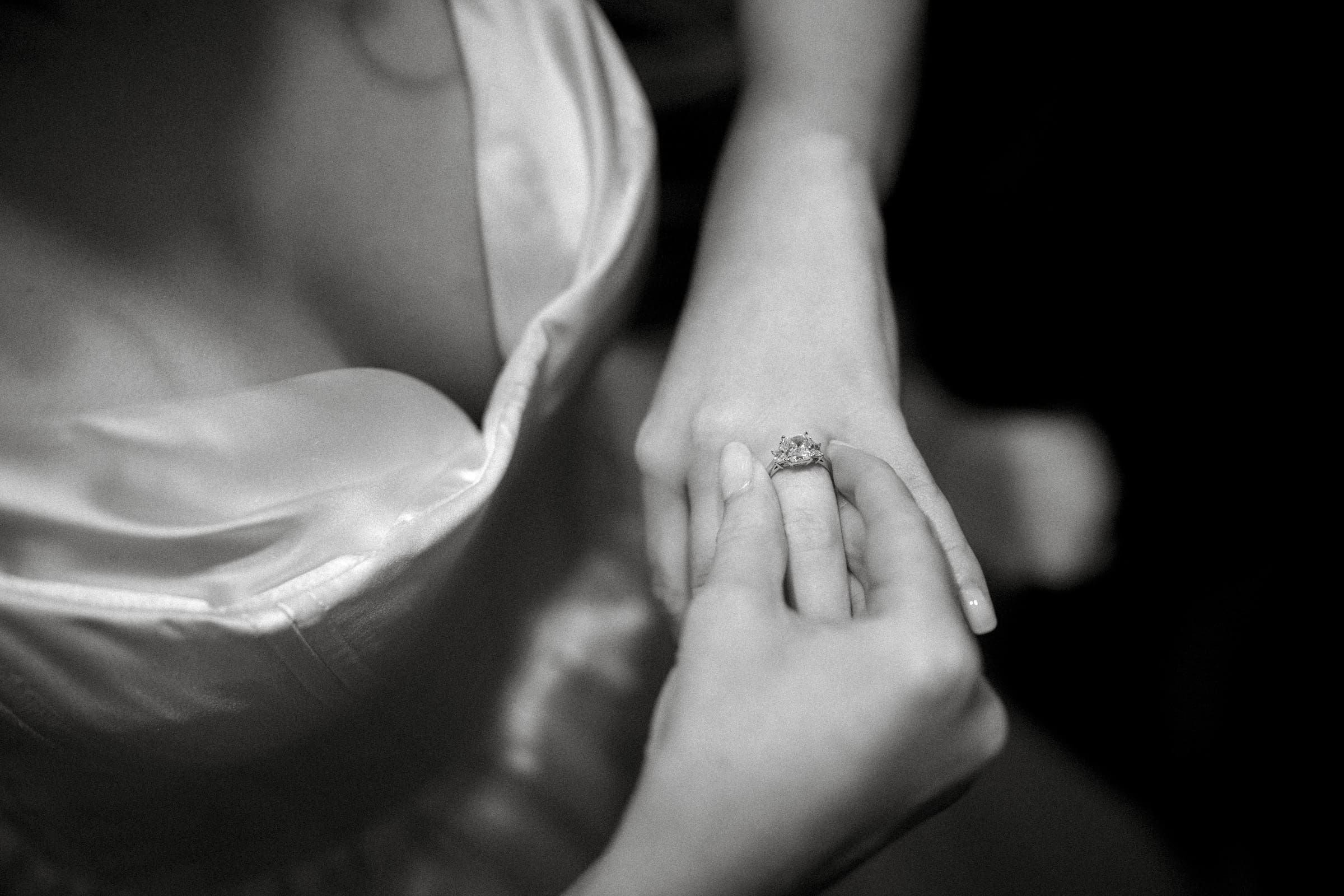 A black and white close-up of a person in a satin dress at an Umstead Hotel Wedding, placing an ornate ring on their finger. The focus is on the hands and ring, creating an intimate and elegant atmosphere.