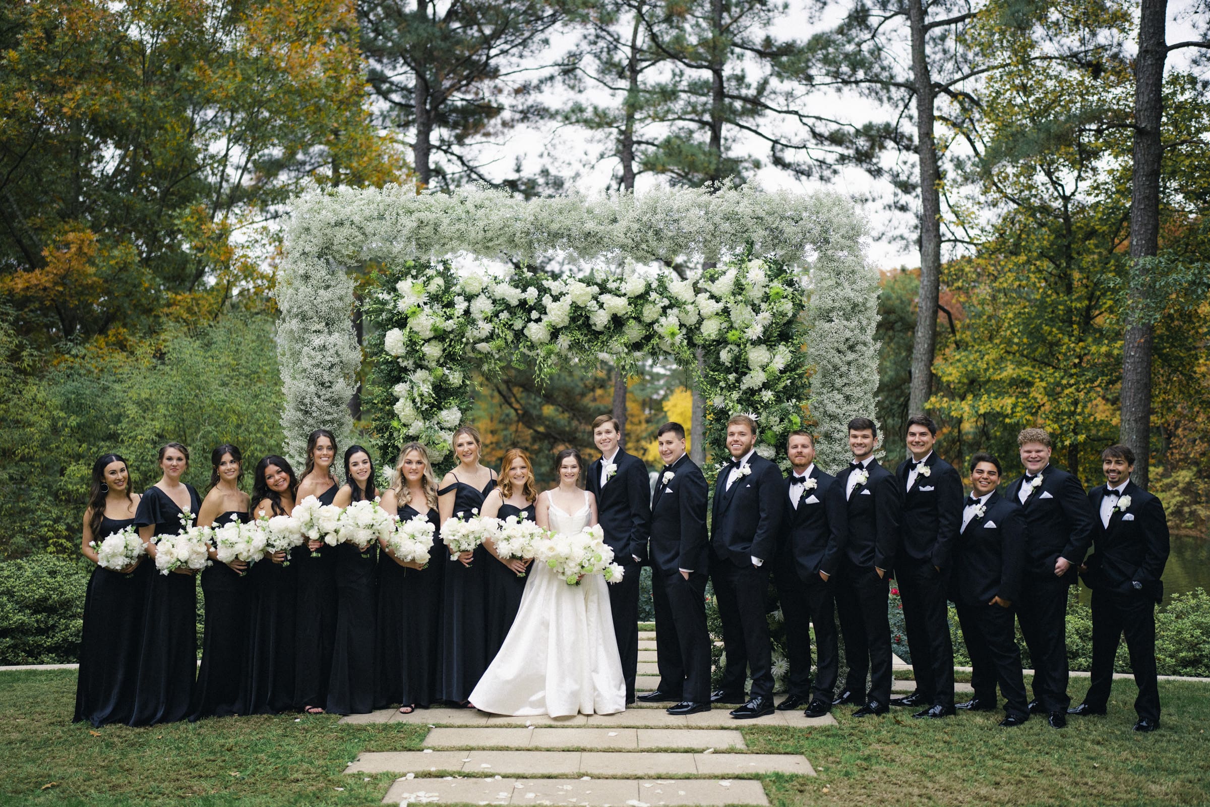 At their Umstead Hotel wedding, the bride and groom stand with their wedding party under a floral arch. The bridal party, in black attire, holds white bouquets. The groomsmen wear black suits and bow ties, all surrounded by lush trees and greenery.