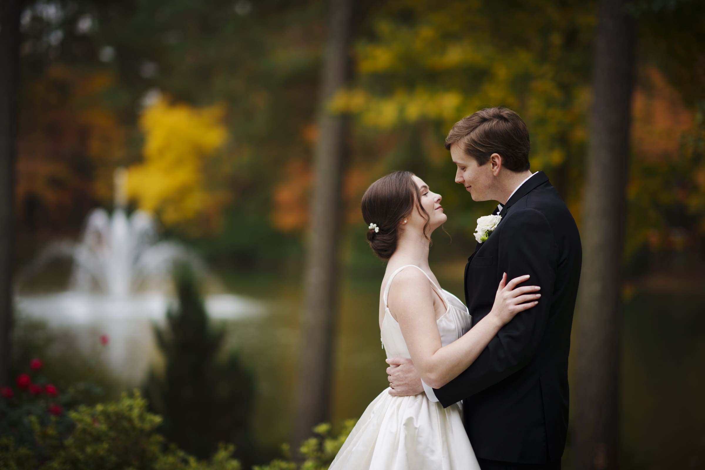 The bride and groom embrace lovingly outdoors by a pond at their enchanting Umstead Hotel wedding, with a large fountain and vibrant autumn trees in the background. She wears a white dress, while he is elegantly outfitted in a black suit.
