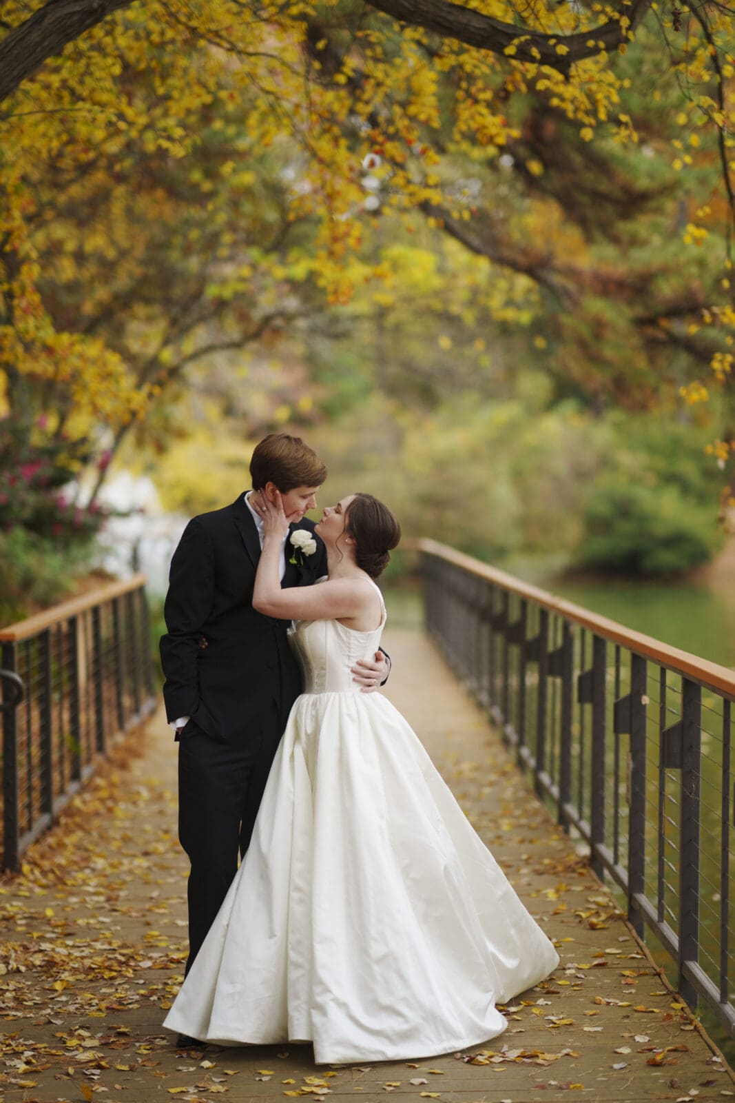 At the scenic Umstead Hotel wedding, a bride and groom kiss on a leafy, tree-lined bridge. The bride wears a white gown, and the groom is in a black suit. Yellow leaves create a vibrant canopy overhead, enhancing the romantic scene.
