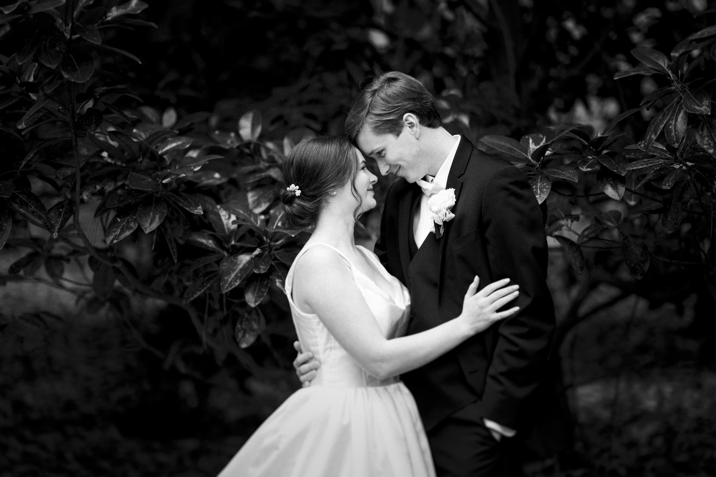 A black and white photo captures a couple embracing in front of leafy trees at their Umstead Hotel wedding. The woman wears a sleeveless gown with a floral hair accessory, while the man dons a suit with a boutonniere. They lean in close, touching foreheads and smiling.