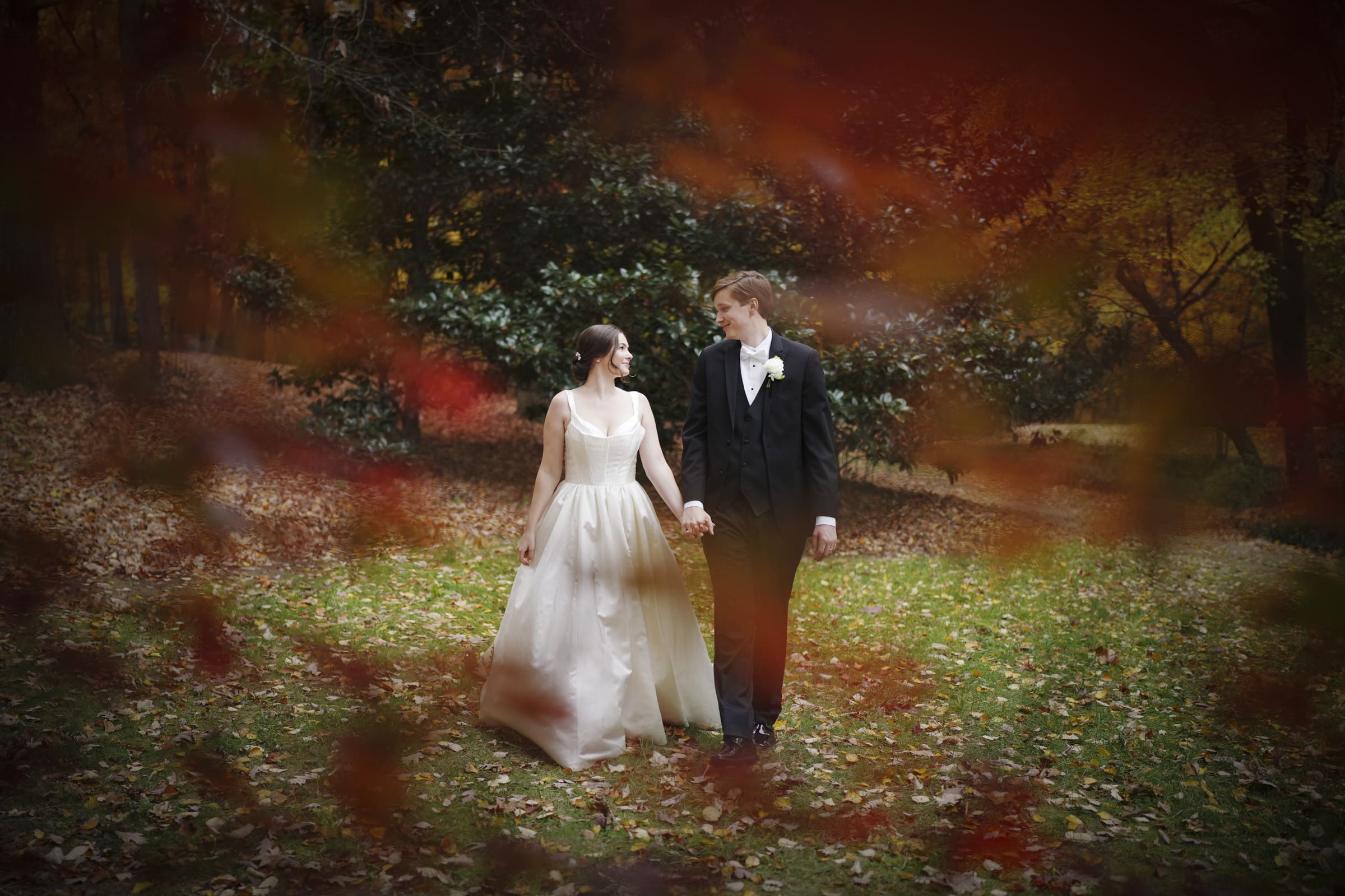 A bride and groom walk hand in hand through a leafy park, reminiscent of an Umstead Hotel wedding. The bride wears a white dress, and the groom sports a black suit with a bow tie. Autumn leaves frame the scene in warm tones, capturing their love amid nature's embrace.