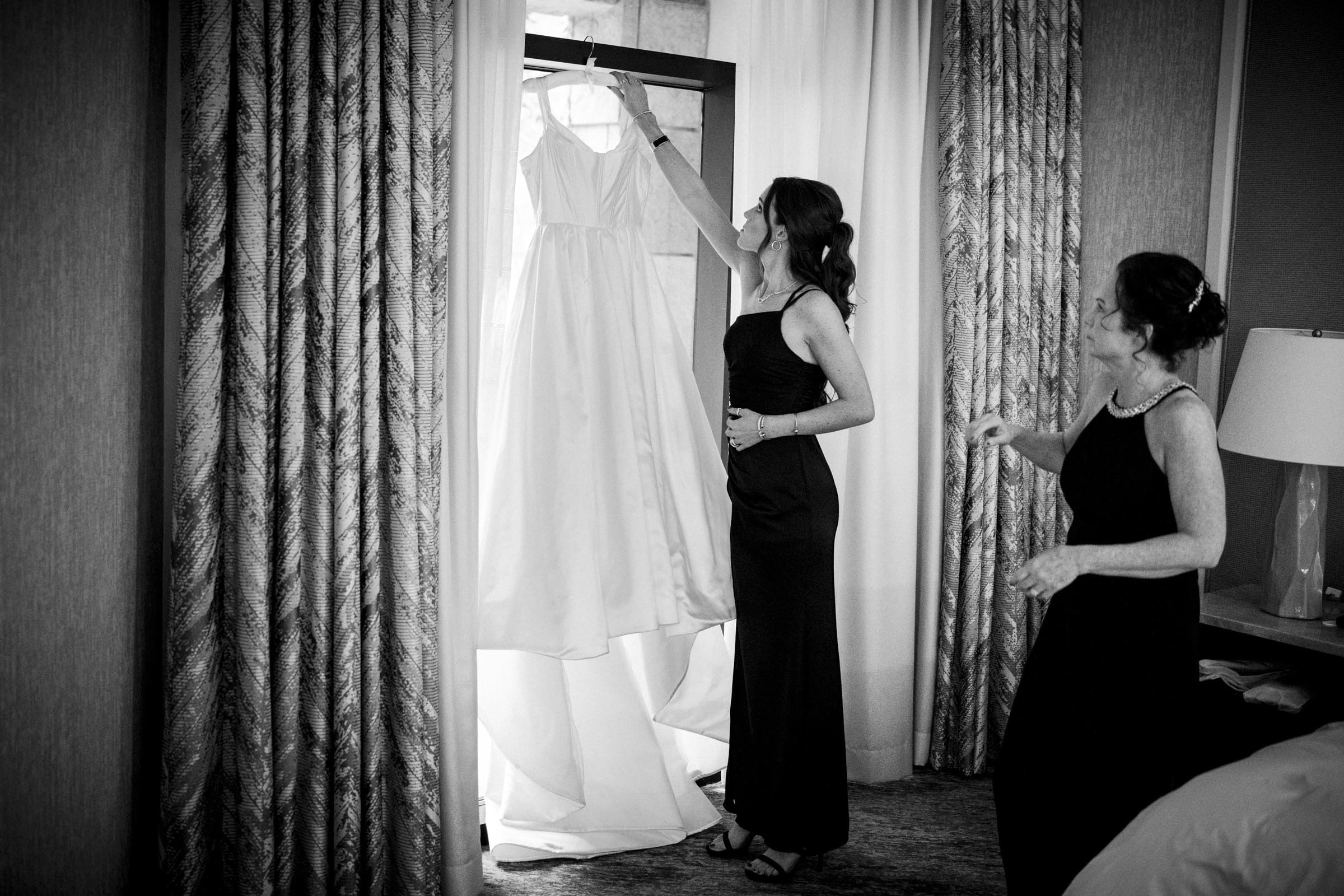 Two women in a softly lit room at the Umstead Hotel admire a wedding dress hanging in front of a window. One holds the dress up, both smiling with anticipation. Curtains frame the window, and a lamp glows gently on the right, setting a serene scene for this special moment.