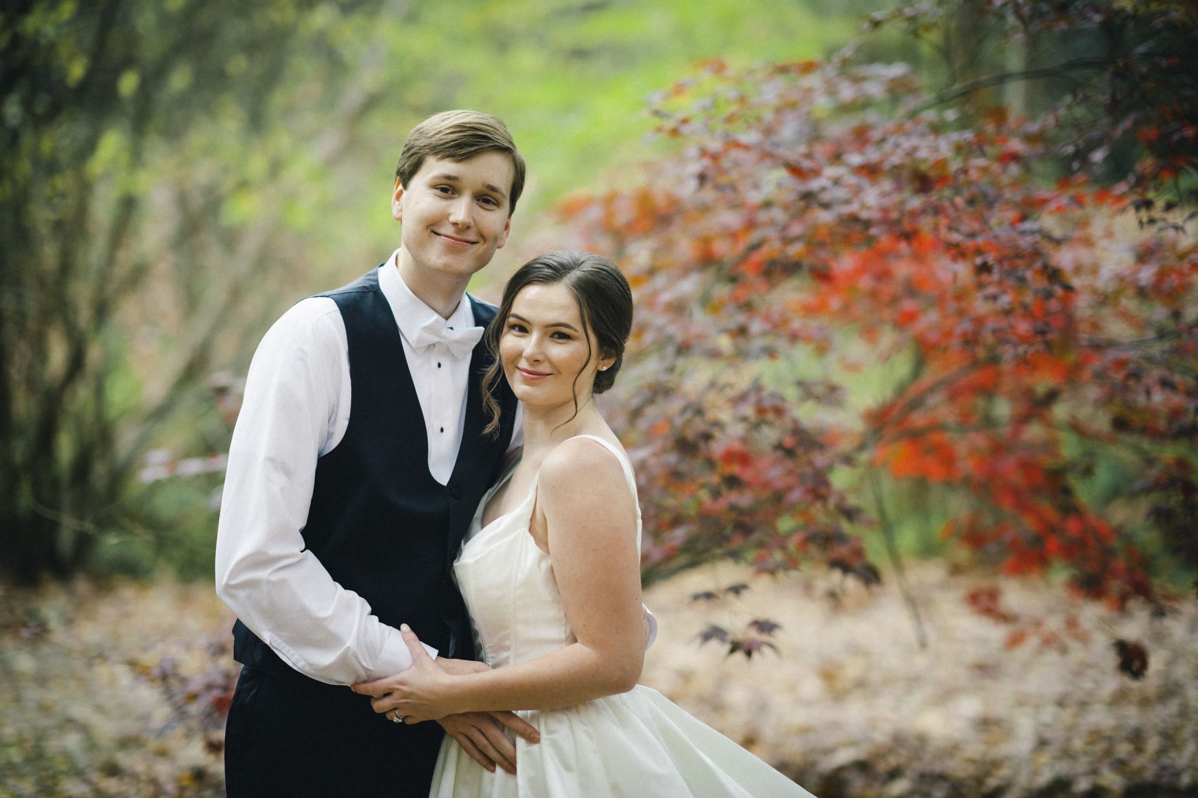 A couple in formal attire stands outdoors, beaming at their Umstead Hotel wedding. The man dons a black vest and bow tie, while the woman shines in a white dress. They are enveloped by leafy trees with vibrant red foliage as their stunning backdrop.
