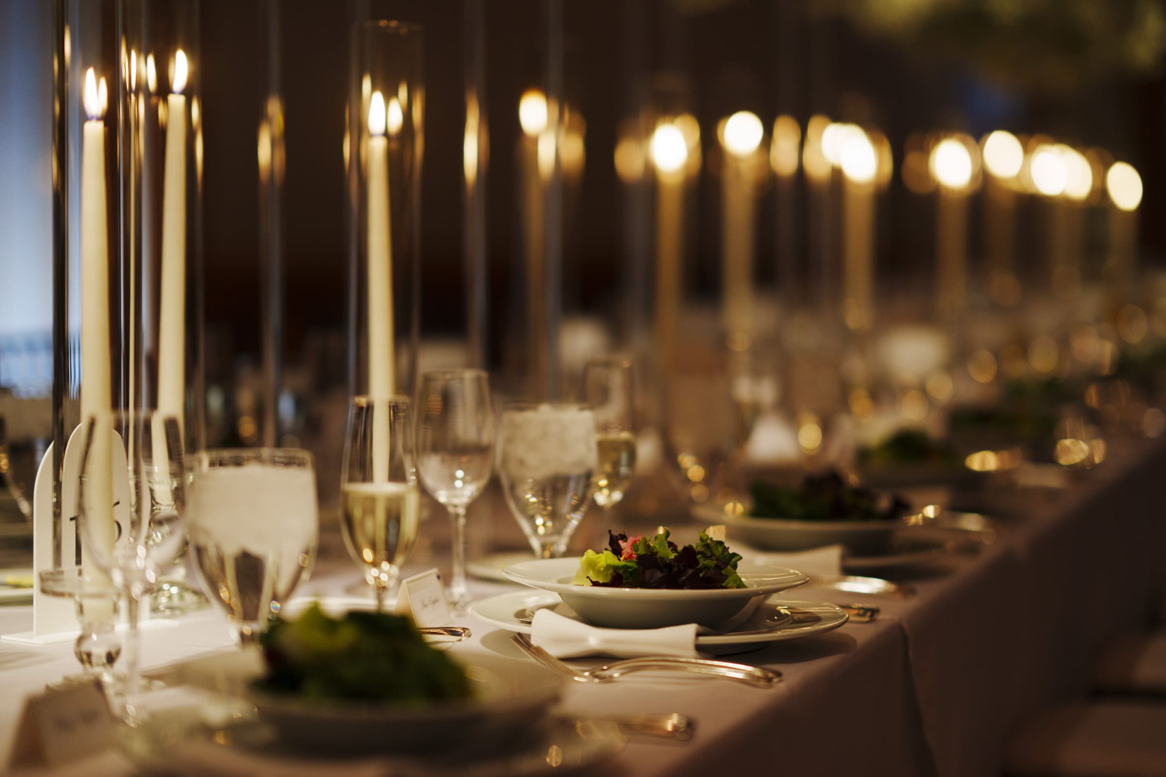 A long, elegantly set dining table at an Umstead Hotel wedding features white tablecloths, plates of salad, clear glassware, and tall, lit candles. The candles create a warm, ambient glow, reflecting a formal and sophisticated atmosphere.