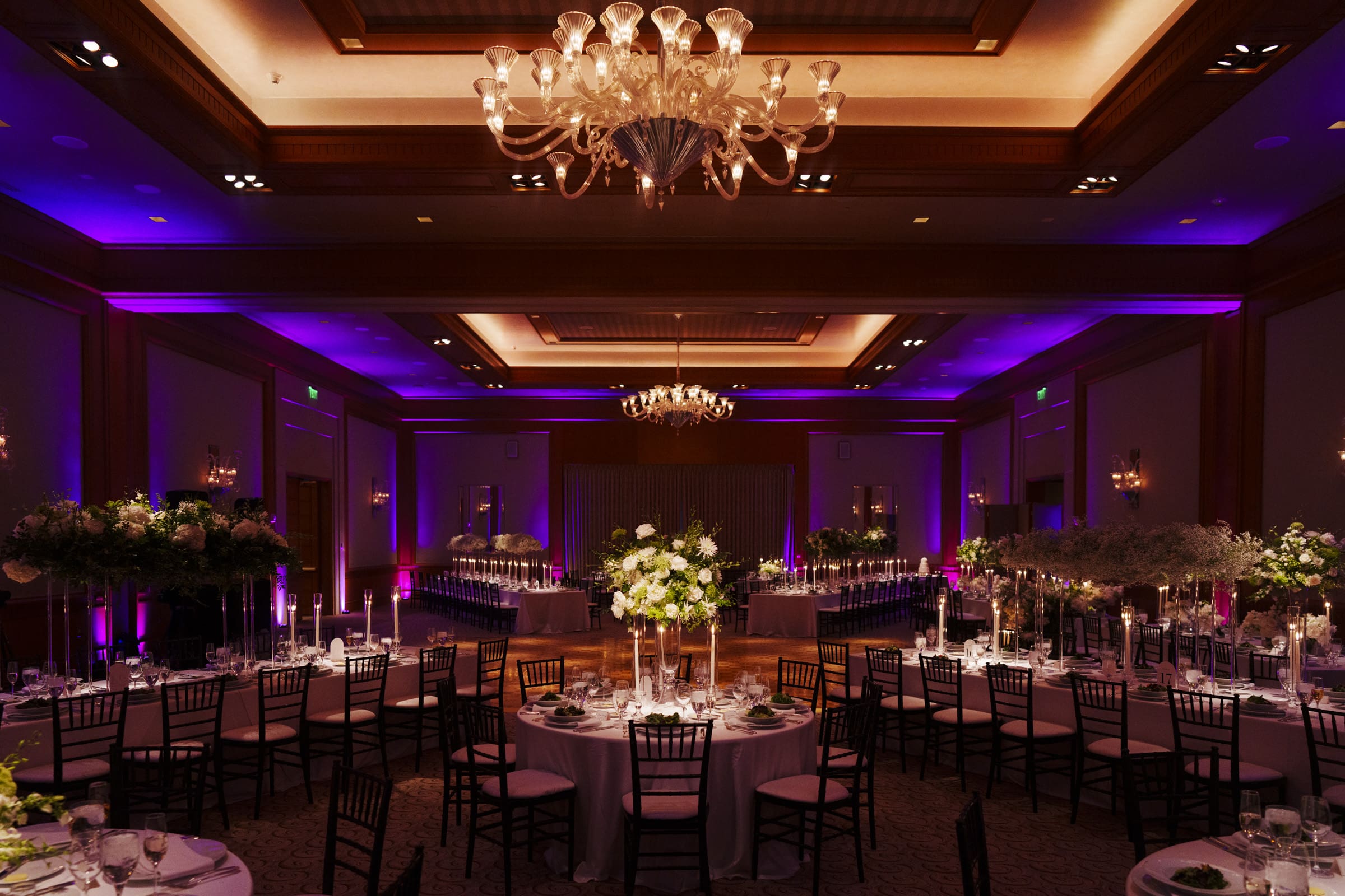 A ballroom at the Umstead Hotel elegantly set up for a wedding reception, featuring round tables with white tablecloths, floral centerpieces, and dark chairs. The room is illuminated with chandeliers and purple lighting, creating a warm and inviting atmosphere.