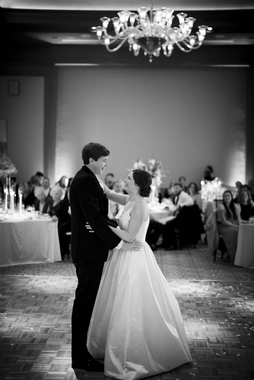 At an Umstead Hotel wedding, a bride and groom share their first dance amid elegantly decorated tables and flickering candles. Guests watch from around the dance floor as a chandelier casts a warm glow over the couple.