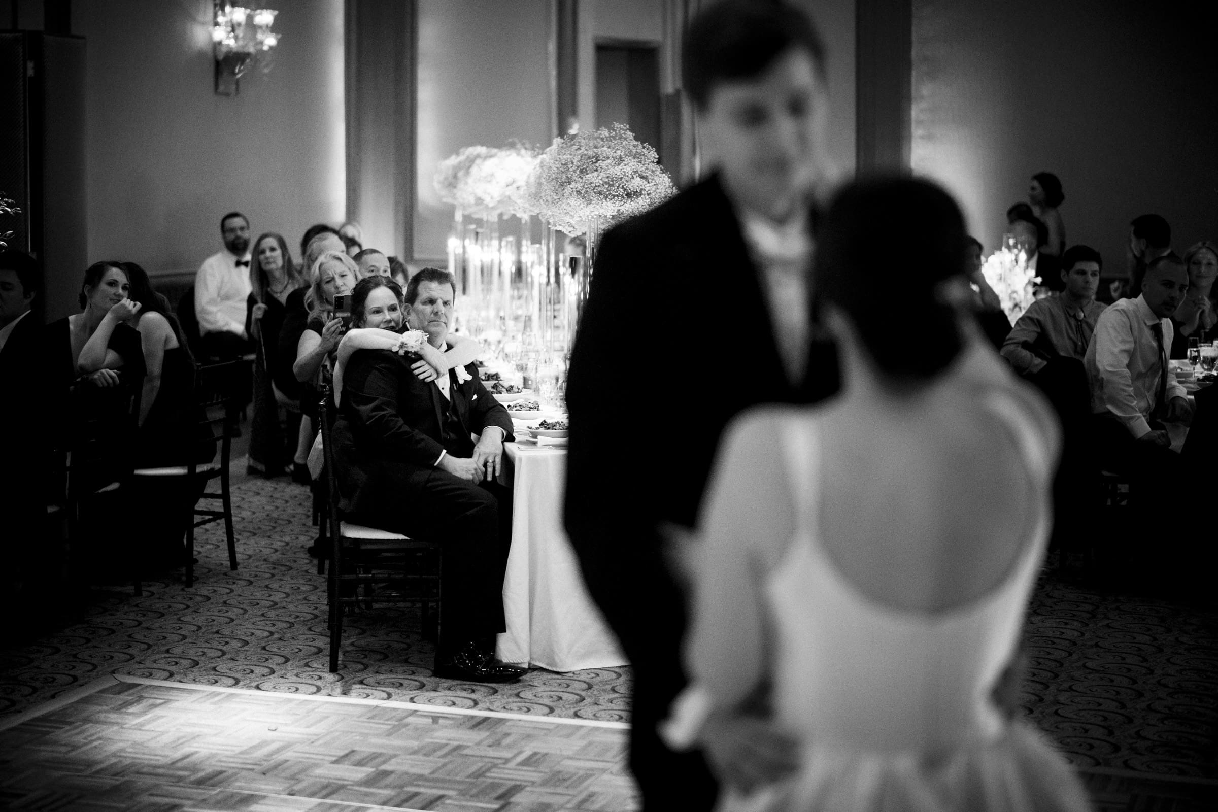 A black and white photo captures a couple dancing gracefully at an Umstead Hotel wedding reception. In the background, guests seated at tables share warm embraces. The room exudes elegance, adorned with candles and exquisite flower arrangements.