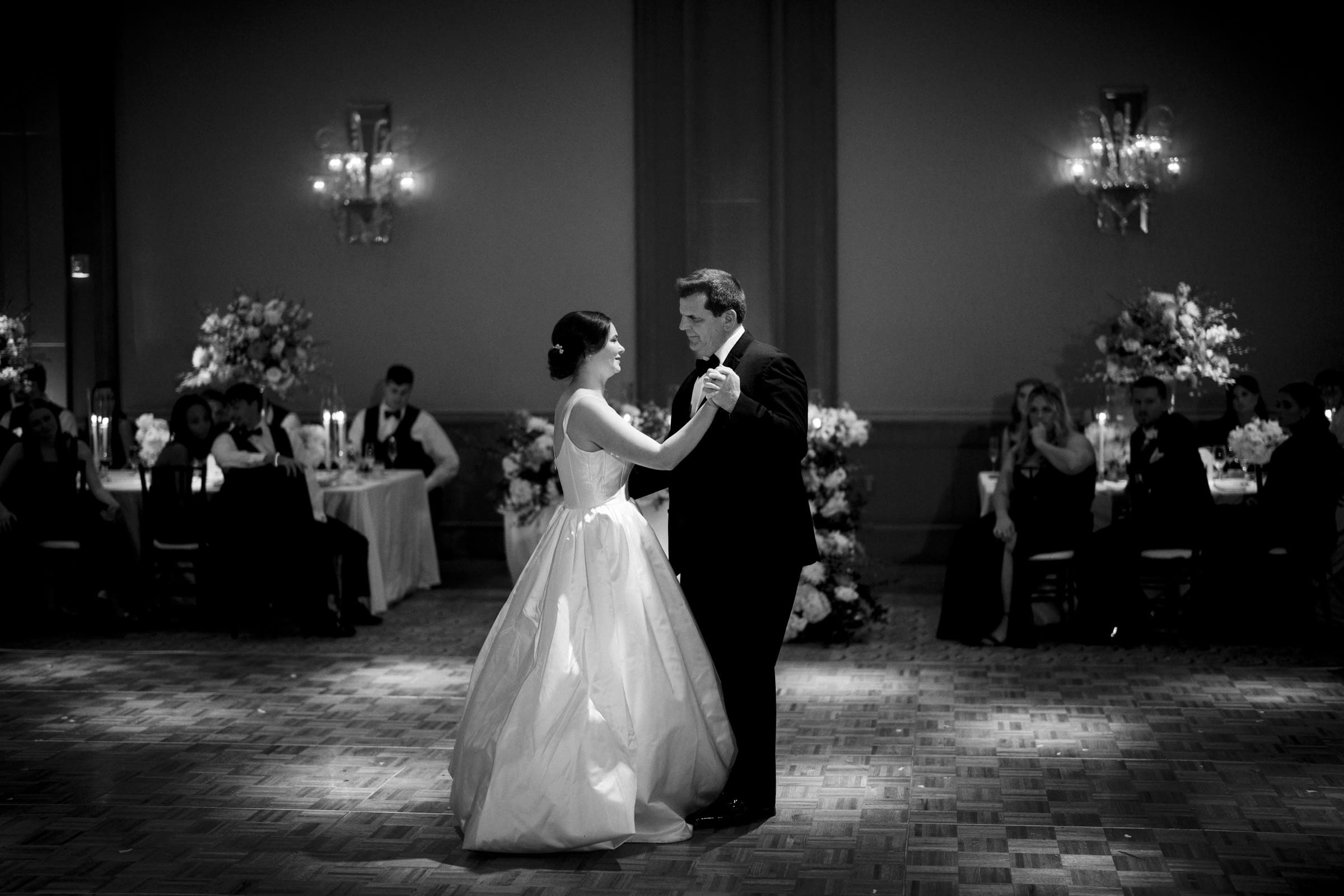A bride and groom share their first dance in a dimly lit reception hall at the Umstead Hotel Wedding. The bride wears a flowing gown, and the groom is in a suit. Guests sit at tables in the background, watching the couple.