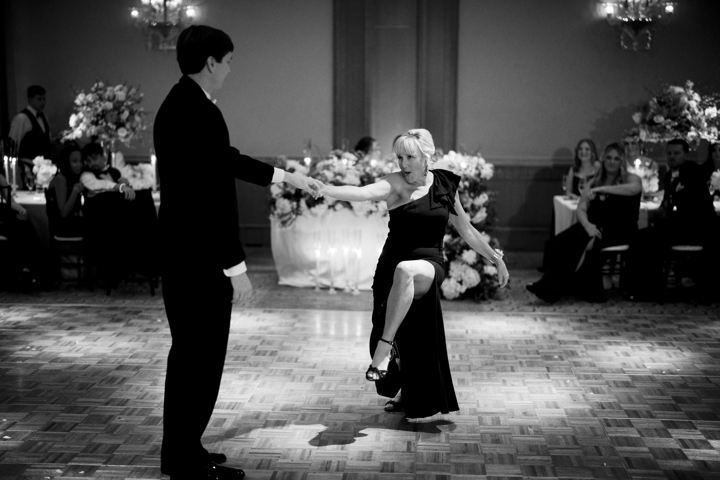 A black and white photo captures a couple dancing gracefully on the wooden floor at an Umstead Hotel wedding. The woman in a dark dress elegantly extends a leg as the man in a suit holds her hand. Guests watch with admiration, surrounded by exquisite floral arrangements.
