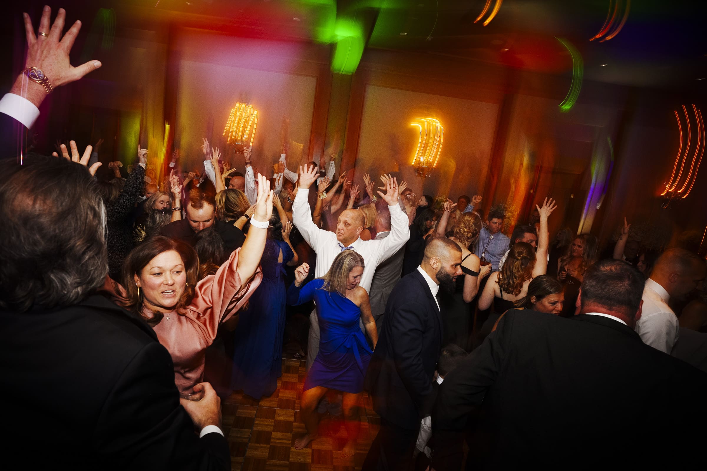 A lively party scene unfolds at an Umstead Hotel wedding, with people dancing energetically on the dance floor. The crowd has their hands raised, and colorful lights illuminate the room, creating a vibrant atmosphere.