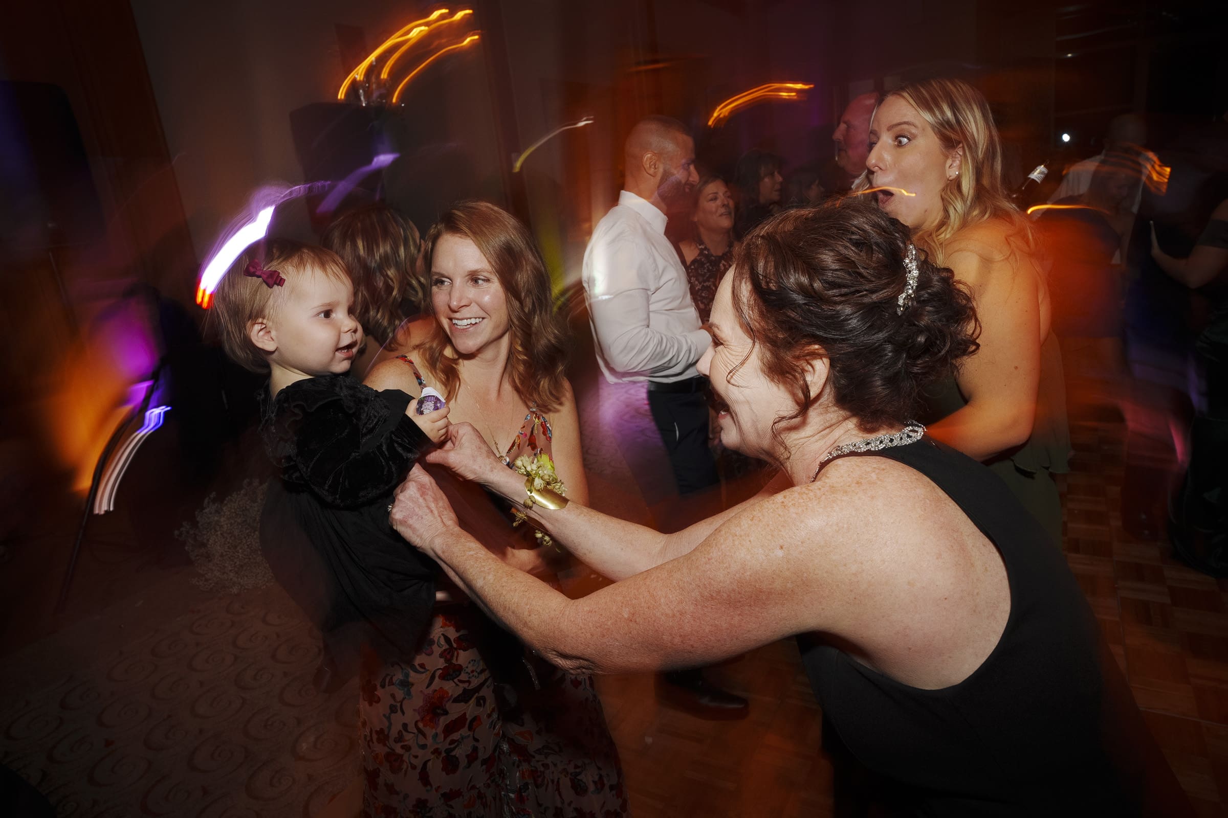 A lively indoor party scene unfolds at an Umstead Hotel wedding, with people dancing joyfully. A woman smiles as she holds a child in her arms, while another woman reaches out to them. Colorful lights create a dynamic, festive atmosphere.
