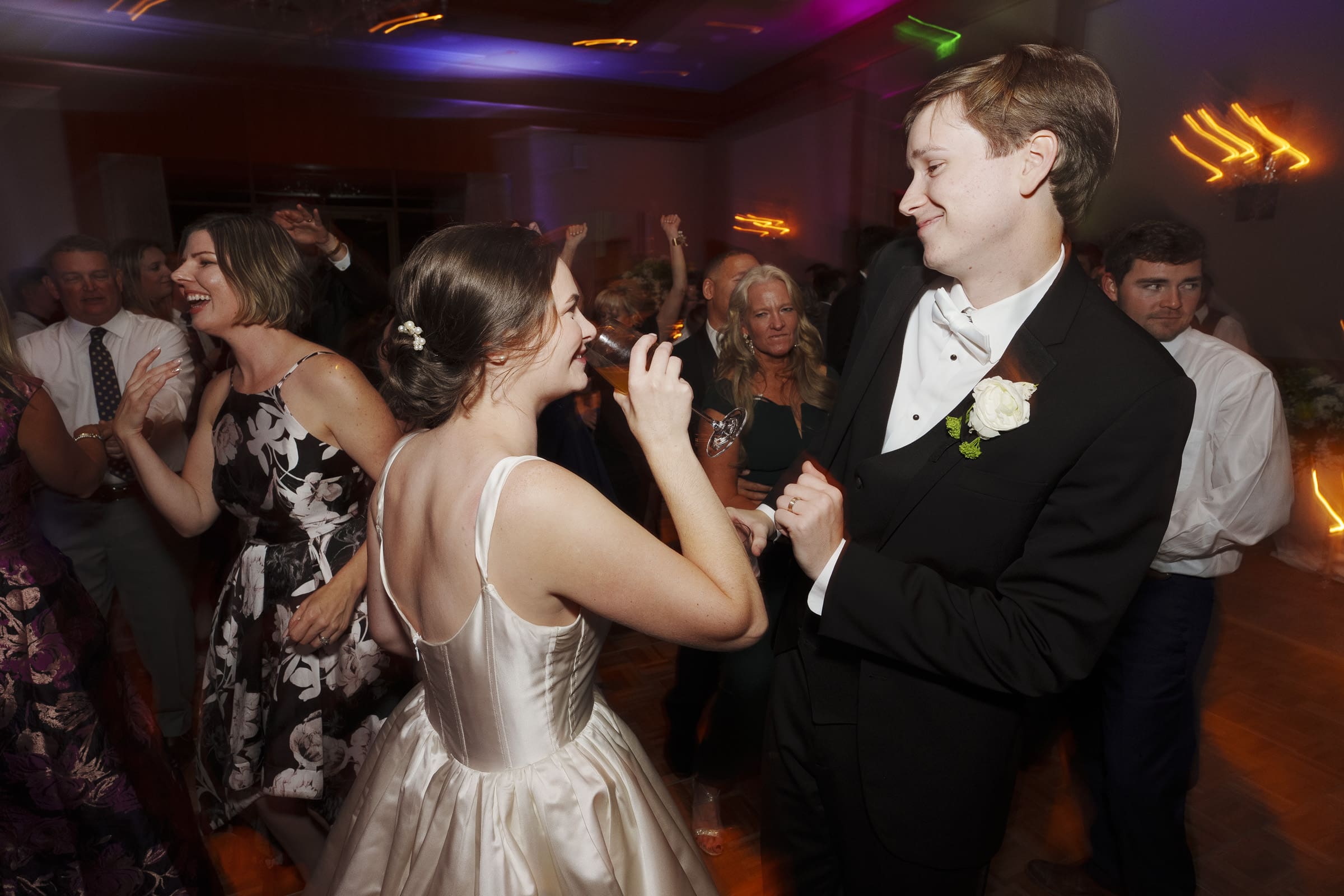 A bride and groom are smiling and dancing at their Umstead Hotel wedding reception. The bride dazzles in a satin dress, while the groom is sharp in a tuxedo. Guests in formal attire surround them, enjoying the celebration under colorful lights.