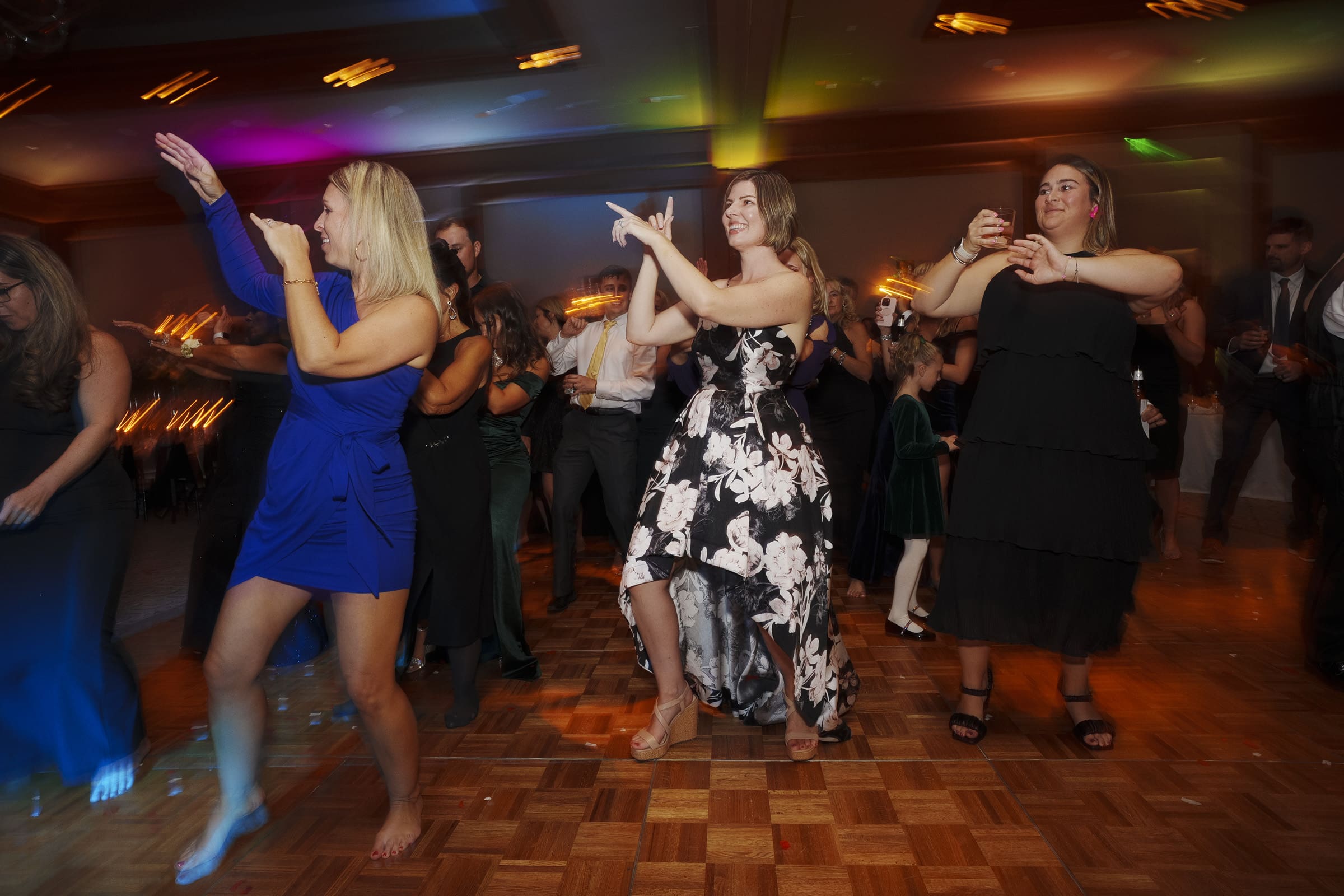 Amidst colorful lights, a lively group dances energetically on the wooden floor. In the front, women twirl in blue and floral dresses, capturing the vibrant spirit of an Umstead Hotel Wedding. Guests in varied attire add to the enchanting celebration unfolding in the dimly lit room.