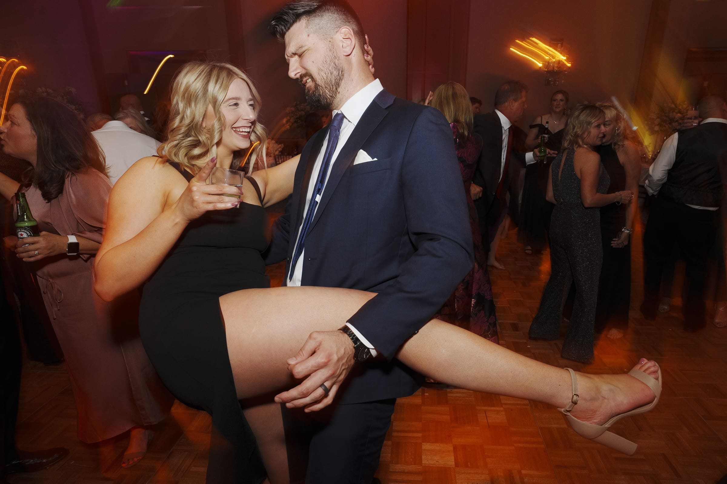 At an Umstead Hotel wedding, a woman in a black dress, holding a drink, playfully lifts her leg while dancing with a man in a suit on the lively dance floor. Others whirl around them, and the atmosphere is festive and joyful.