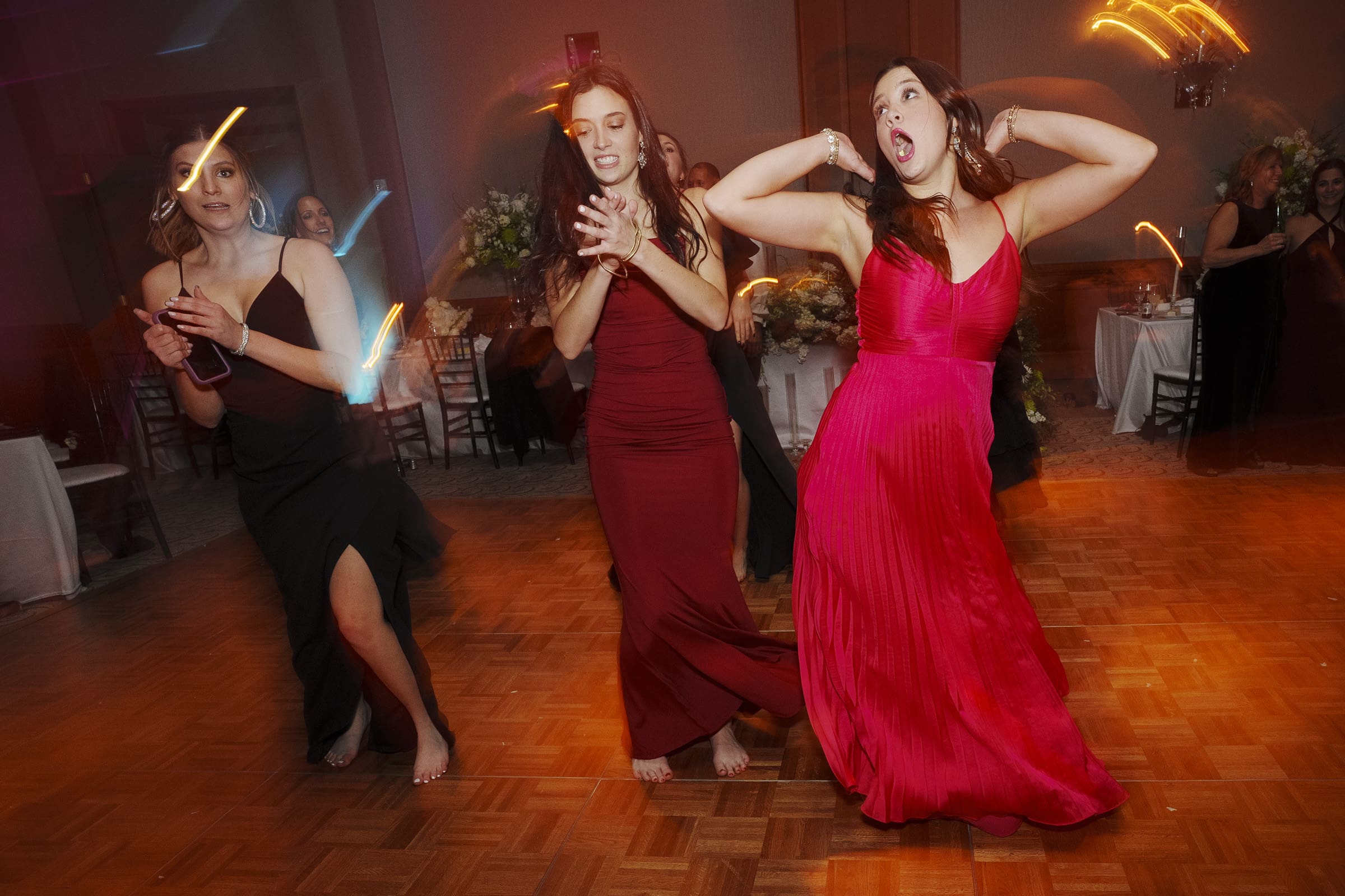 At an elegant Umstead Hotel wedding, three women in formal dresses dance energetically on a wooden floor. One wears a black dress, another a burgundy dress, and the third a bright pink dress. The room is dim, and light streaks add a dynamic effect to the lively scene.