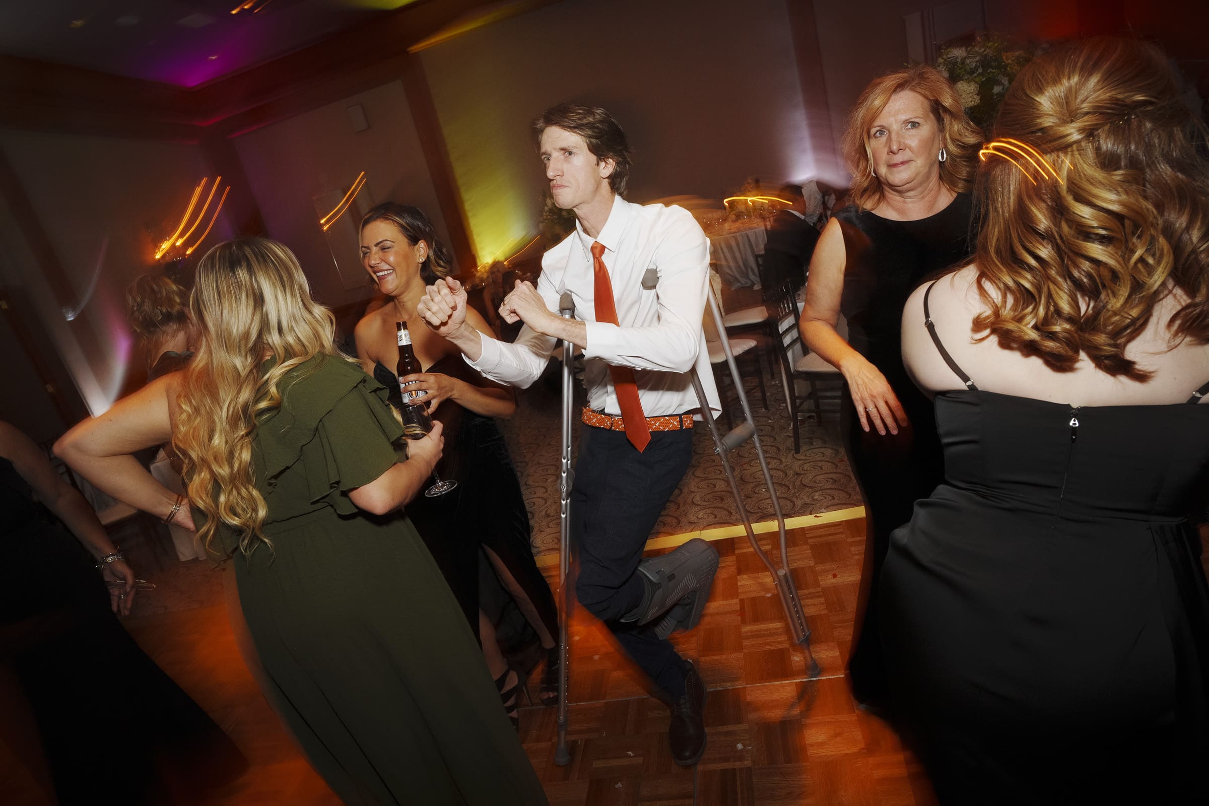 A lively dance floor scene at an Umstead Hotel wedding shows a man with crutches enthusiastically dancing with several women in formal attire. The background is softly lit, creating a warm and celebratory atmosphere.