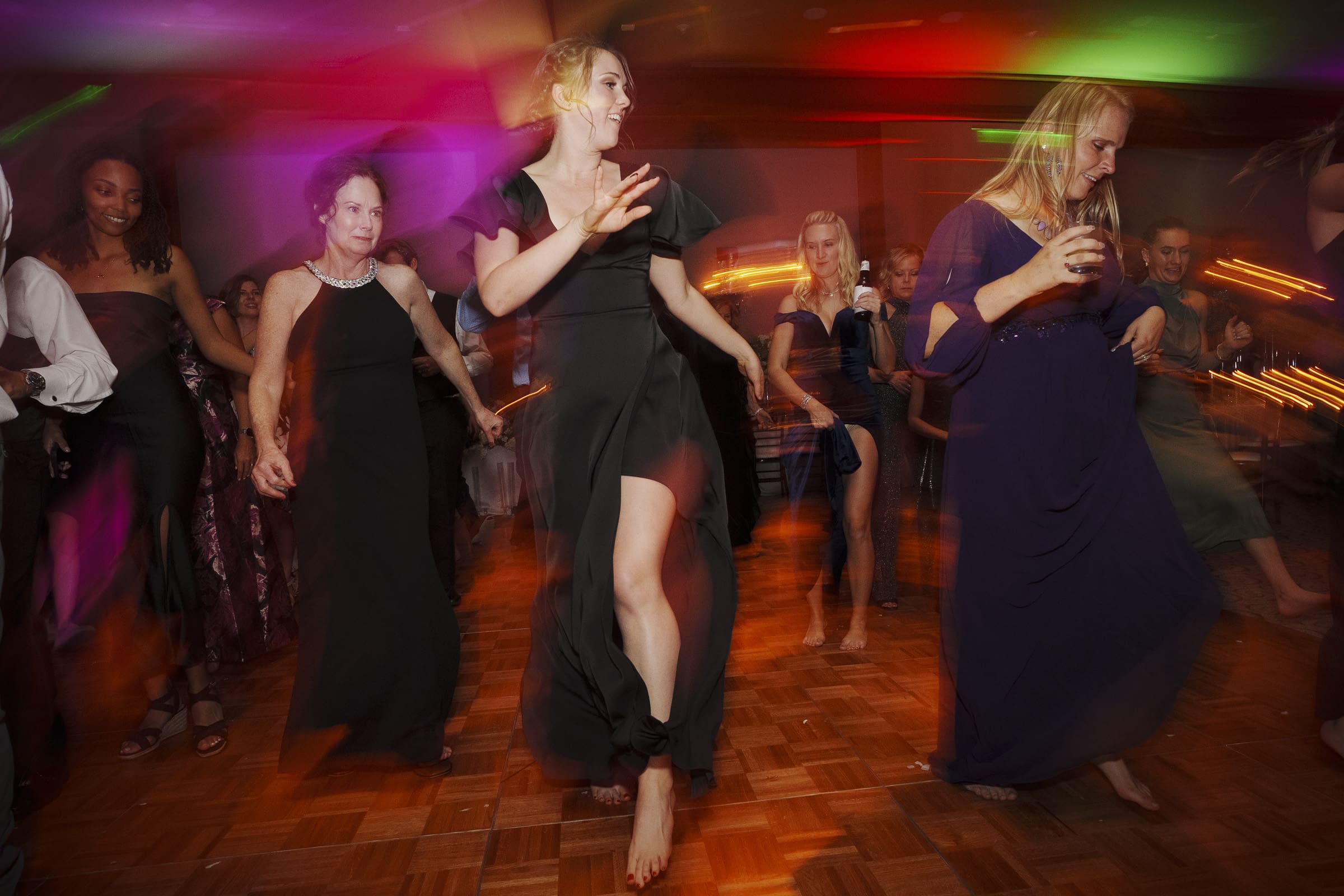 A lively group of people dancing barefoot on a wooden dance floor at an Umstead Hotel wedding, dressed in formal and semi-formal attire. Colorful lights illuminate the room, creating a festive atmosphere.