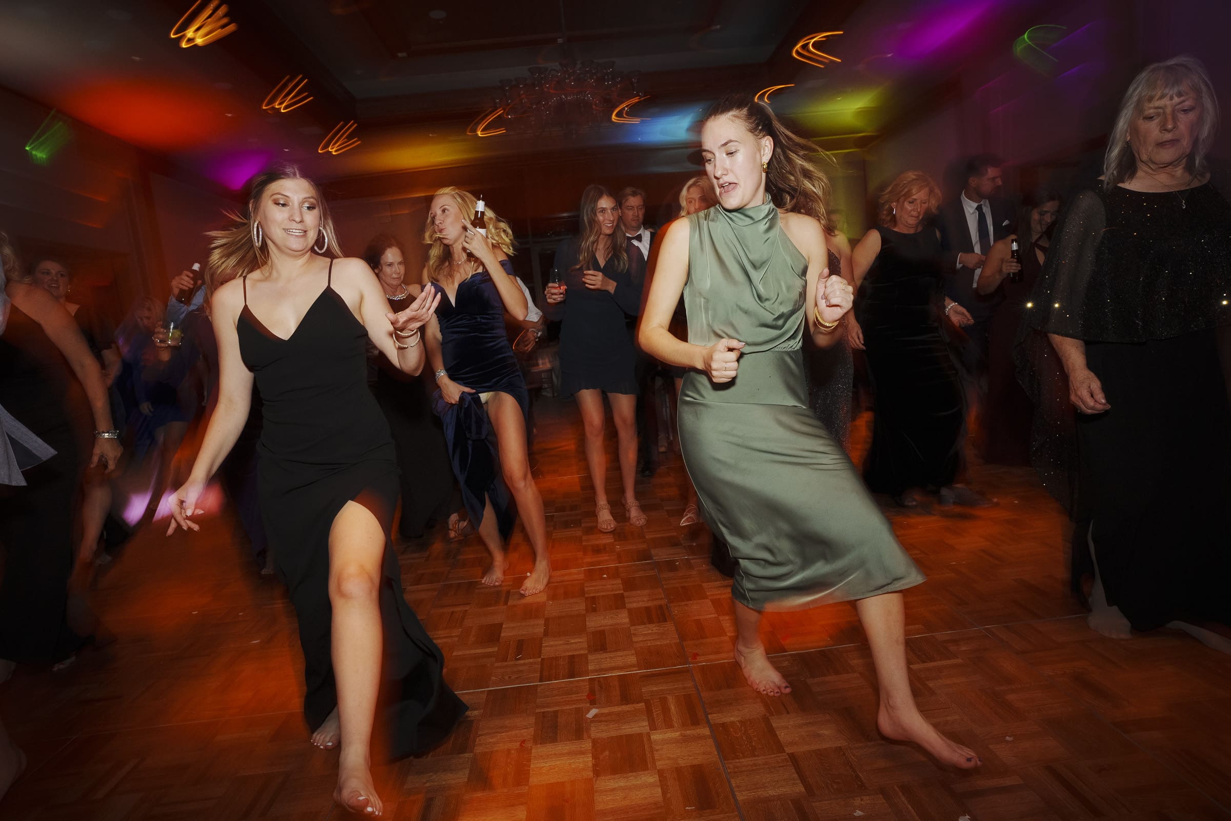 A lively Umstead Hotel wedding scene with people dancing enthusiastically on a wooden floor. The room is illuminated by colorful lights, and everyone appears to be enjoying the vibrant atmosphere. Some women wear elegant dresses as they dance energetically.