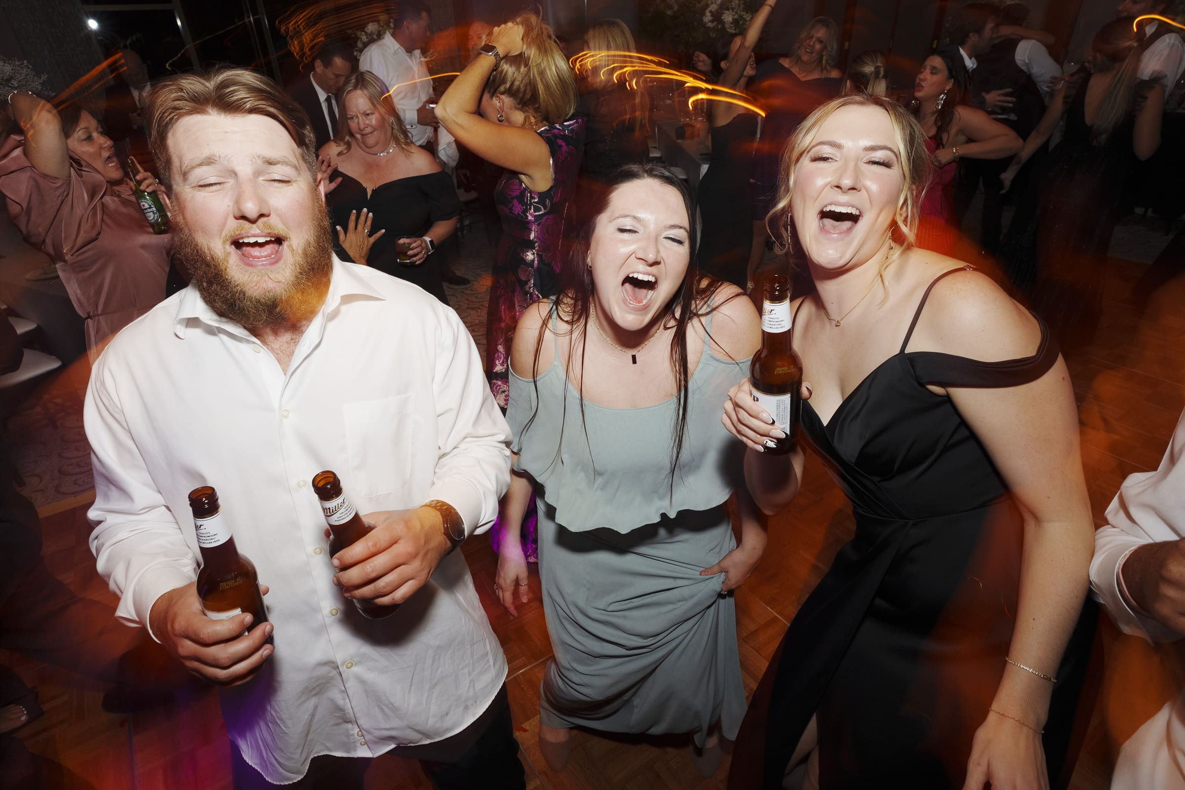 Three people holding drinks, laughing, and enjoying themselves at a lively indoor party, much like an Umstead Hotel wedding. The scene is full of movement with blurred lights and other guests dancing in the background. Everyone appears to be having a great time.